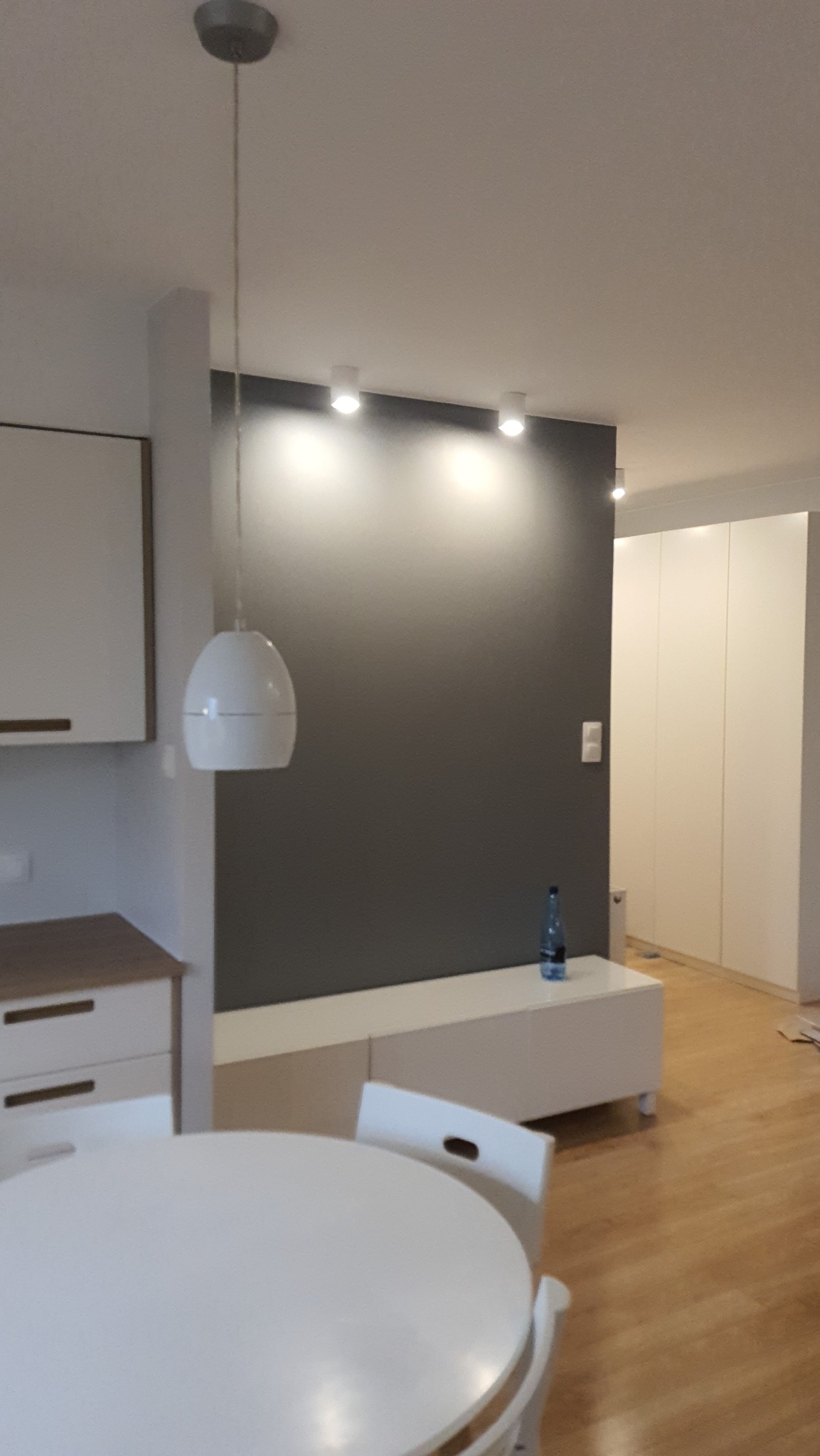 Szara ściana z oświetleniem punktowym w nowoczesnym salonie, biały stół i krzesła na pierwszym planie, butelka wody na białej komodzie, drewniana podłoga.