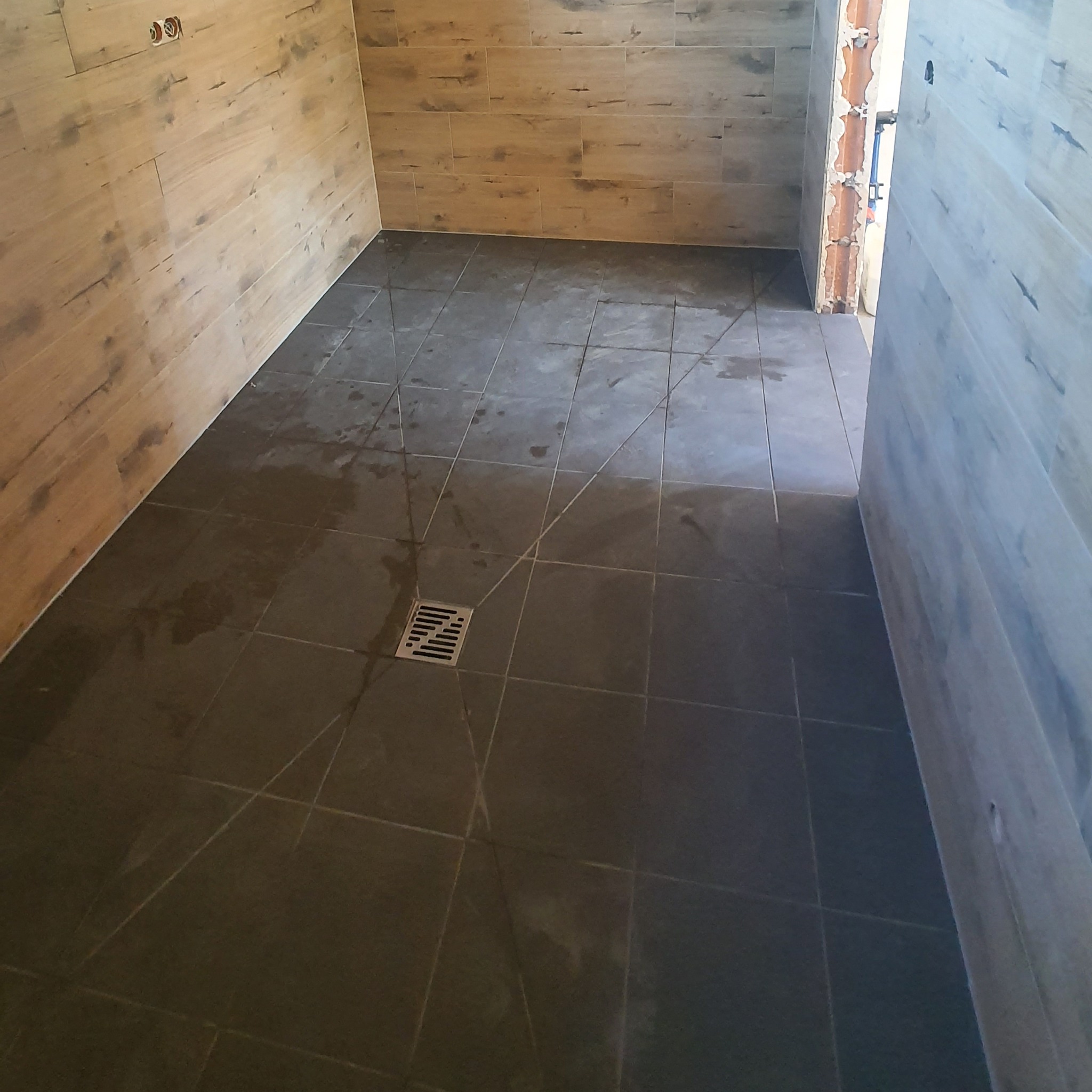 Świeżo wykafelkowana podłoga w łazience z odpływem liniowym, na ścianach płytki imitujące drewno, widoczne ślady po fugowaniu.