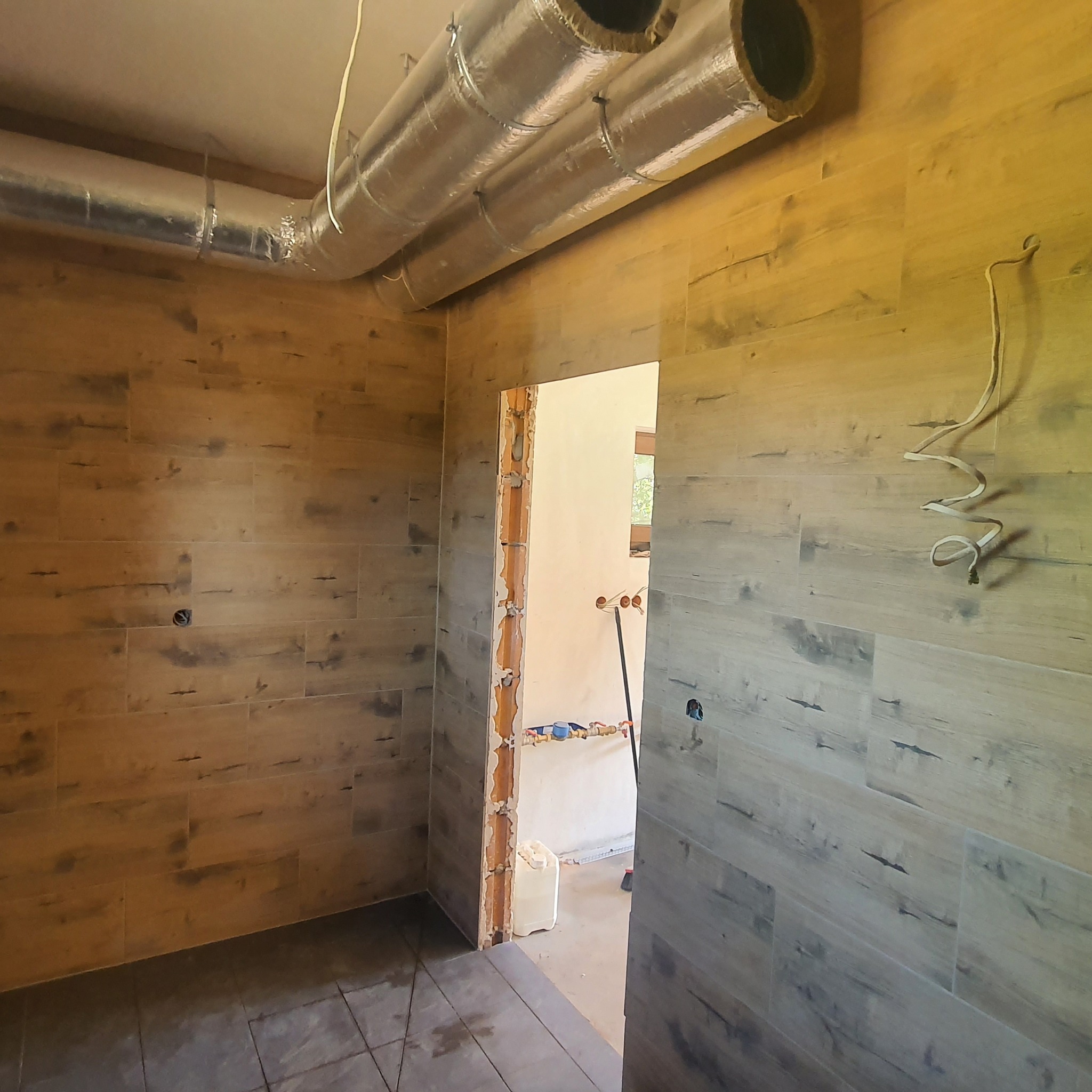 Wnętrze łazienki w trakcie remontu: ściany wyłożone płytkami imitującymi drewno, widoczne fragmenty instalacji wentylacyjnej pod sufitem, otwór drzwiowy z odsłoniętą cegłą i instalacjami...