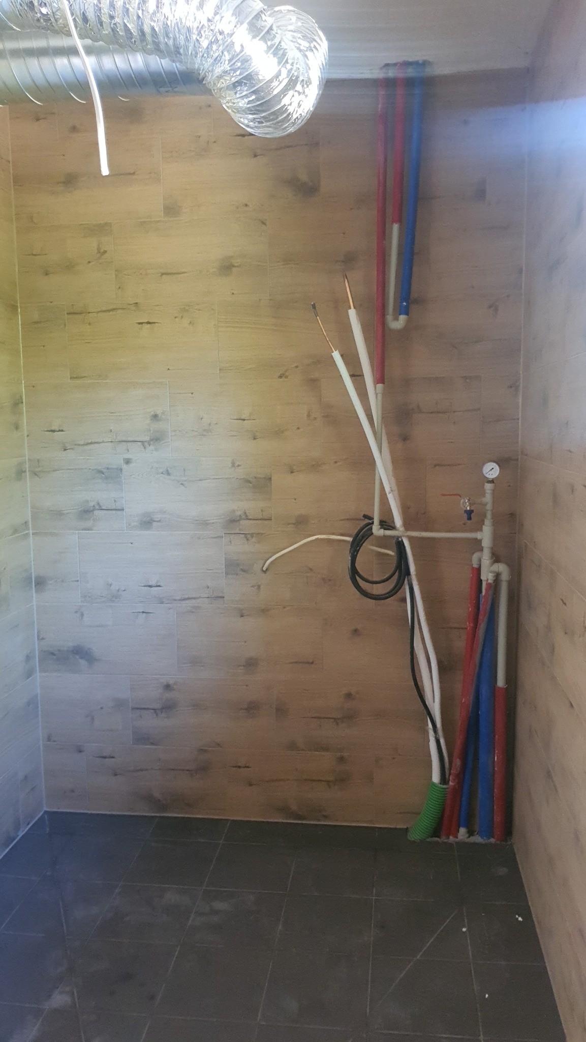 Wnętrze łazienki w trakcie prac instalacyjnych: widoczne rury wodne w kolorach czerwonym i niebieskim, przewody elektryczne, fragment instalacji wentylacyjnej pod sufitem oraz wykończone płytki...