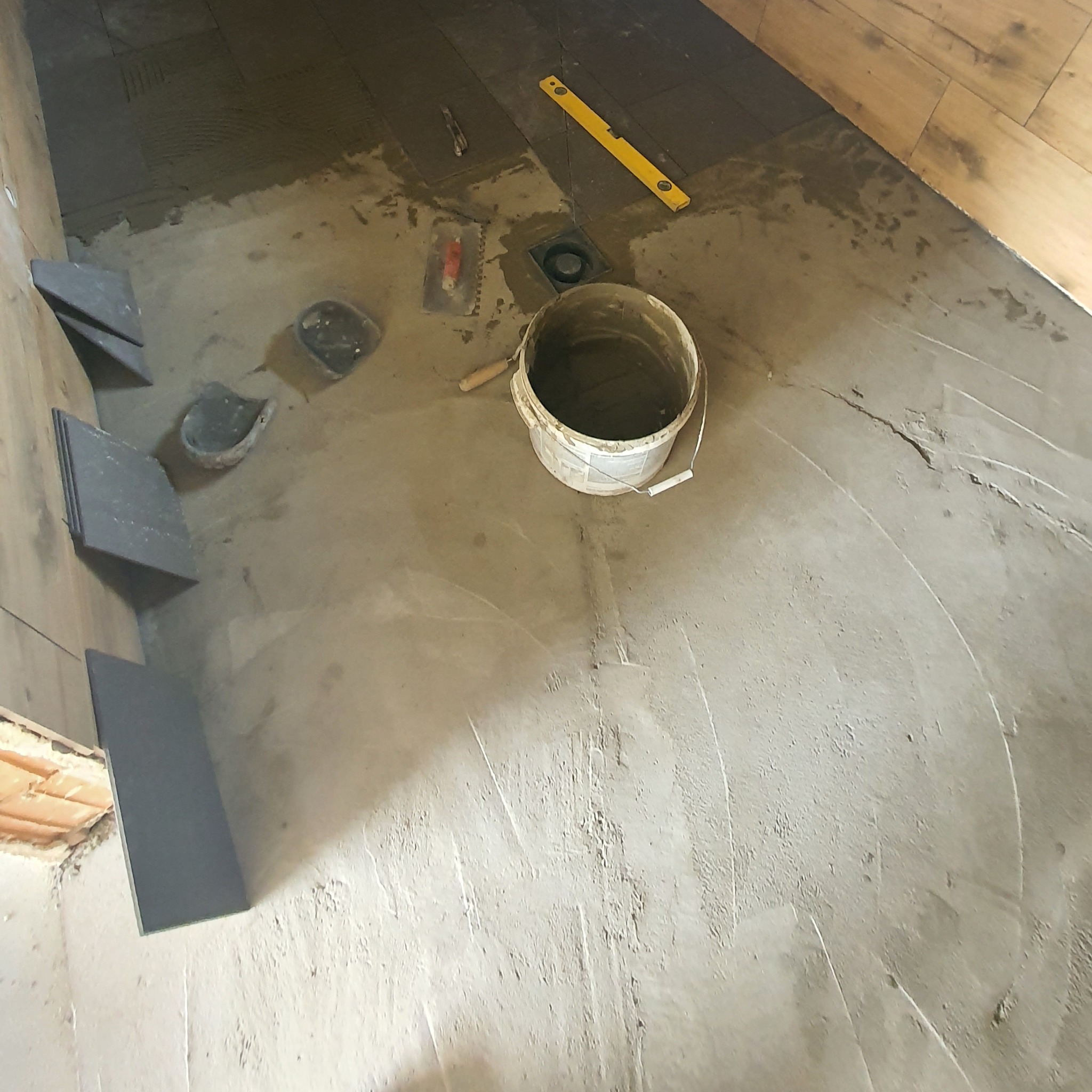 Przygotowanie podłoża pod płytki w łazience, widoczne narzędzia, poziomica, wiadro z zaprawą i ułożone płytki przy ścianie.