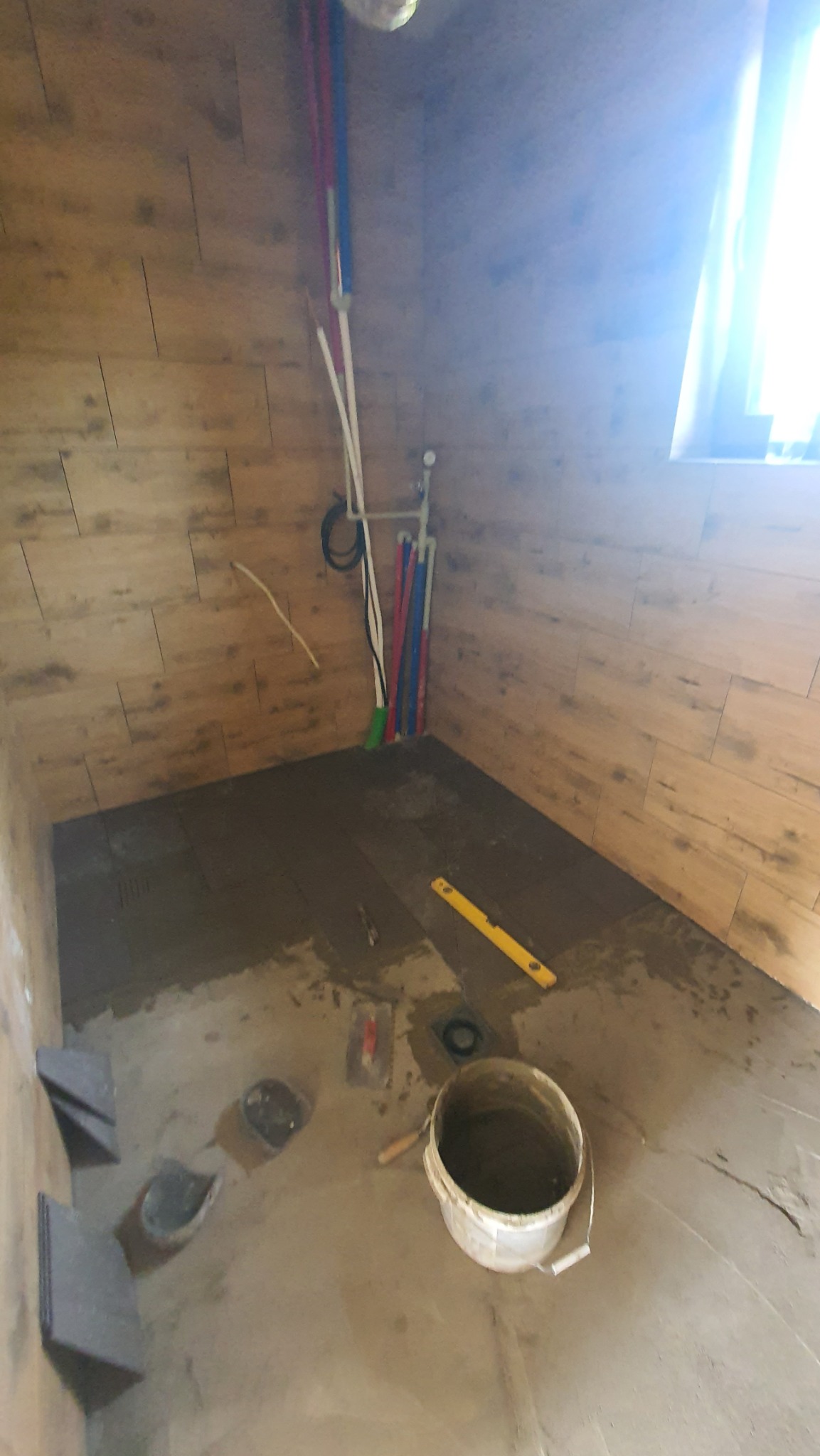 Łazienka w trakcie prac: częściowo ułożone płytki podłogowe, poziomica, wiadro z zaprawą, rury instalacji wodnej i kanalizacyjnej przy ścianie wyłożonej płytkami imitującymi drewno.