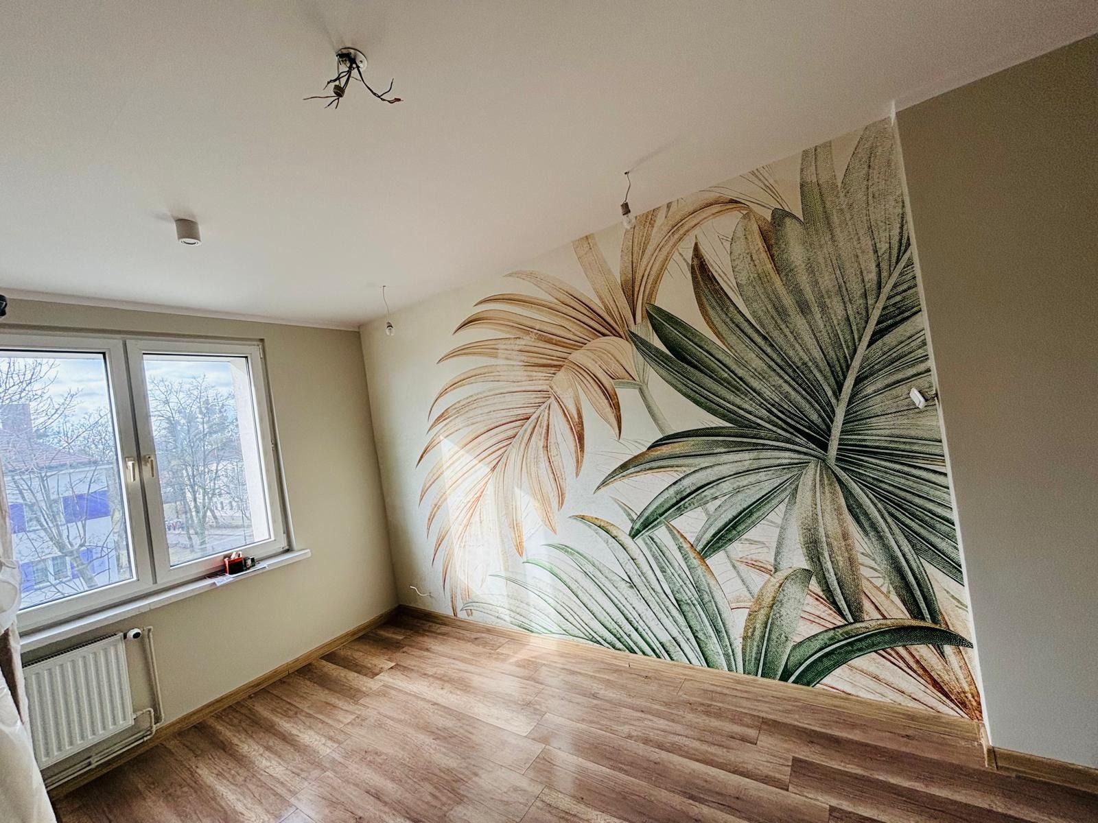 Pokój po remoncie z fototapetą w motywy tropikalne, brązowe panele podłogowe, widoczne kable od oświetlenia sufitowego.