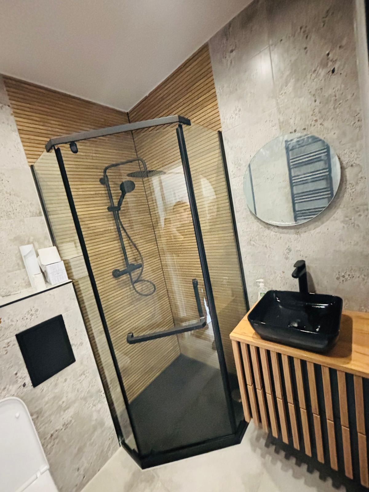 Nowoczesna łazienka z kabiną prysznicową w czarnej ramie, drewnianą boazerią, umywalką nablatową i owalnym lustrem na ścianie z szarego betonu.