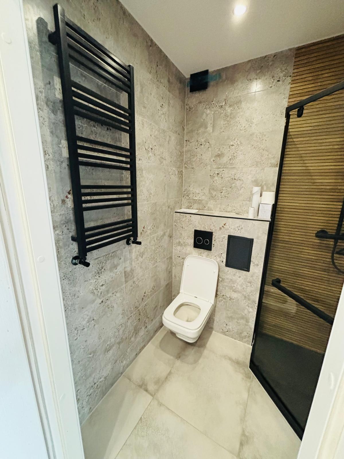 Wykończona łazienka z płytkami imitującymi kamień, czarnym grzejnikiem drabinkowym, toaletą wiszącą i kabiną prysznicową z czarnymi profilami i drewnopodobnymi panelami.