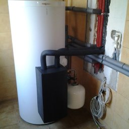 JANUSZ KRAWCZUK CIEPŁOBUDOWA - Biały zasobnik ciepłej wody użytkowej z czarnym modułem grzewczym i niebieskim naczyniem przeponowym, podłączony do instalacji grzewczej z rurami w izolacji czerwonej i szarej, widoczne złącza...