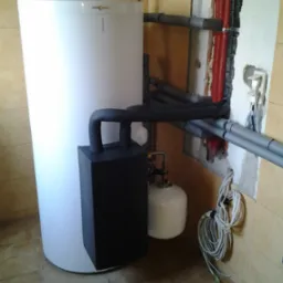 Biały zasobnik ciepłej wody użytkowej z czarnym modułem grzewczym i niebieskim naczyniem przeponowym, podłączony do instalacji grzewczej z rurami w izolacji czerwonej i szarej, widoczne złącza...