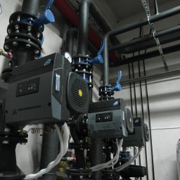 JANUSZ KRAWCZUK CIEPŁOBUDOWA - Rząd zainstalowanych pomp obiegowych Grundfos z manometrami i zaworami odcinającymi, połączonych rurami w kotłowni.