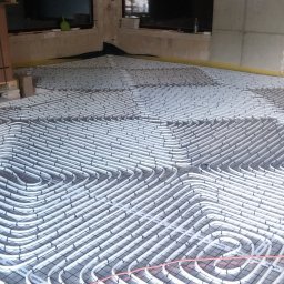 JANUSZ KRAWCZUK CIEPŁOBUDOWA - Białe rury grzewcze Upor ułożone spiralnie na czarnej siatce w surowym pomieszczeniu, widoczny kartonowy kontener marki Viessmann na palecie w tle.