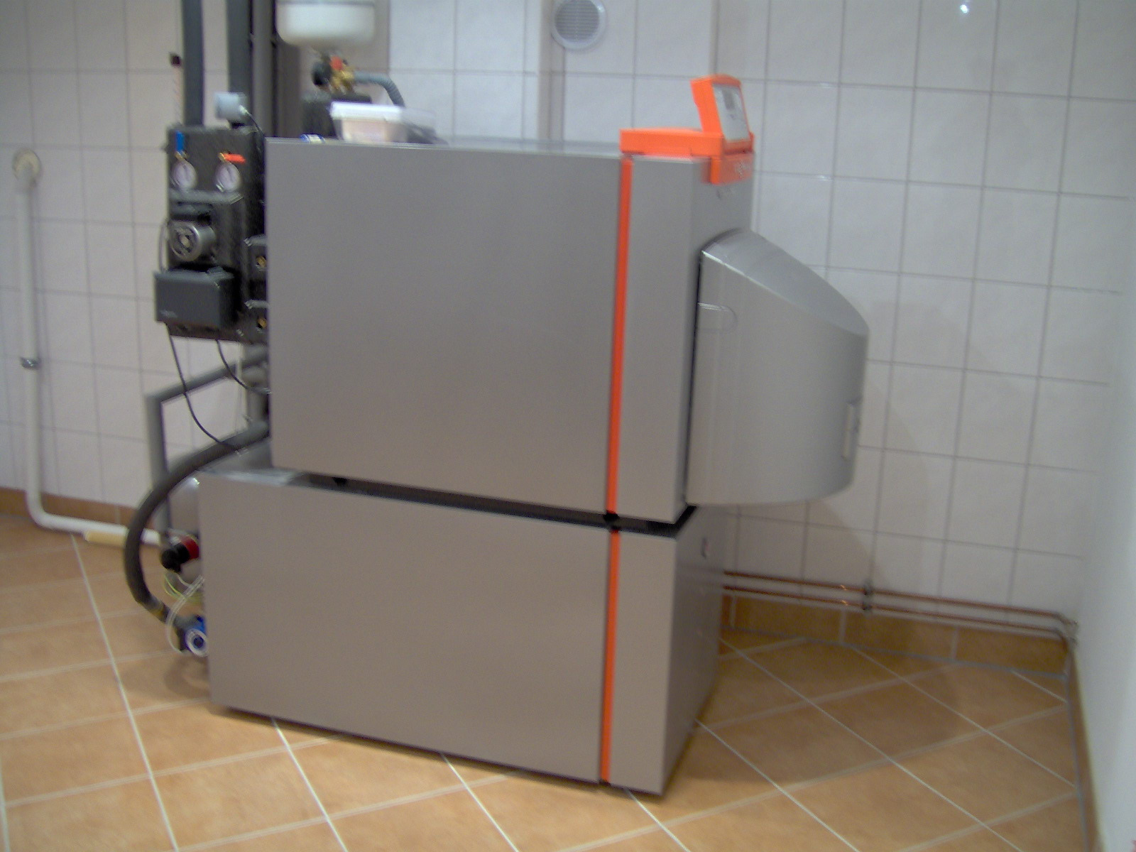 Szary piec na paliwo stałe z pomarańczowymi akcentami, zainstalowany w pomieszczeniu z kafelkami na ścianach i podłodze. Widoczne rury i elementy instalacji.