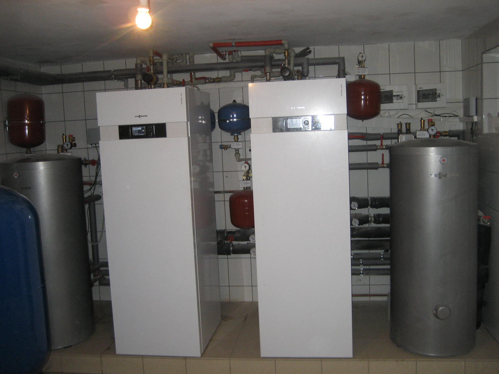 Dwa białe, nowoczesne piece grzewcze marki Viessmann stoją obok srebrnych zbiorników akumulacyjnych w pomieszczeniu kotłowni z białymi płytkami ściennymi i brązowymi płytkami podłogowymi, widoczne...