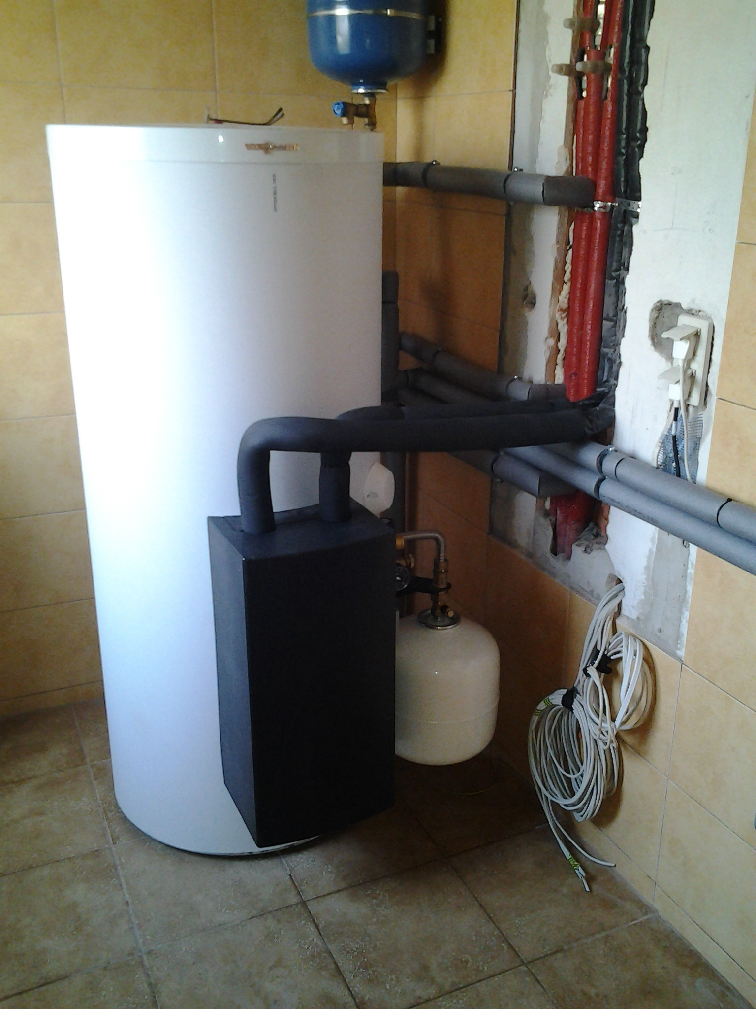 Biały zasobnik ciepłej wody użytkowej z czarnym modułem grzewczym i niebieskim naczyniem przeponowym, podłączony do instalacji grzewczej z rurami w izolacji czerwonej i szarej, widoczne złącza...