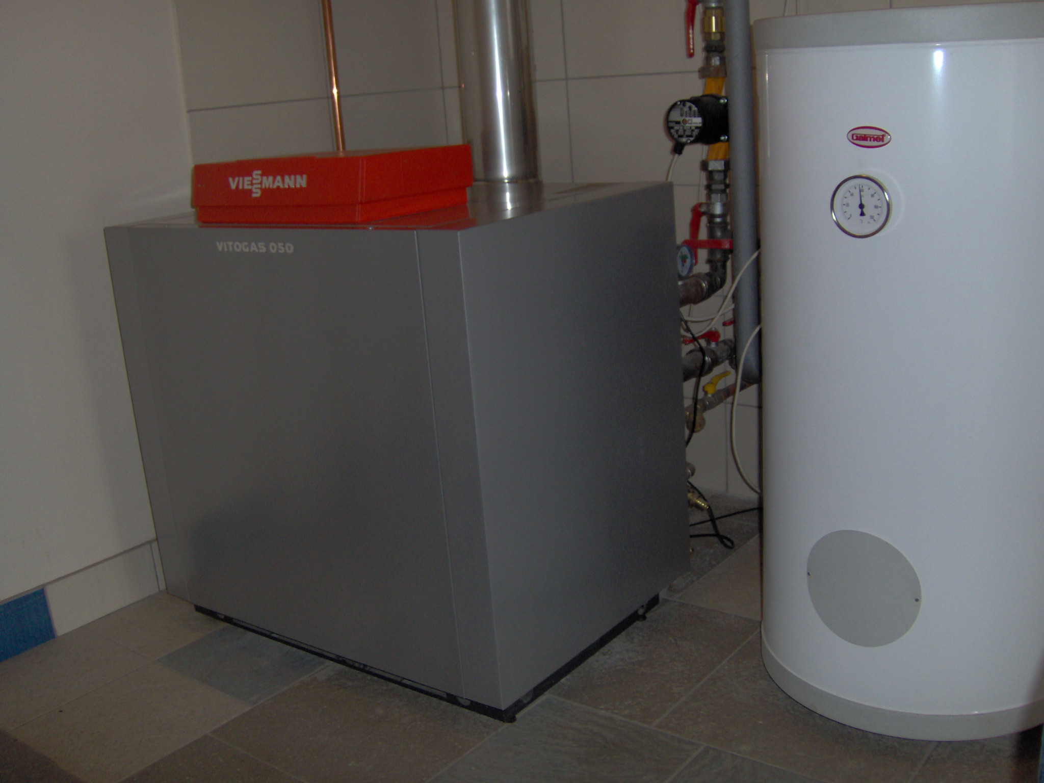 Szary piec gazowy Viessmann Vitogas 050 z czerwonym pudełkiem na górze, obok biały zasobnik ciepłej wody użytkowej Galmet z manometrem, na tle białych płytek.