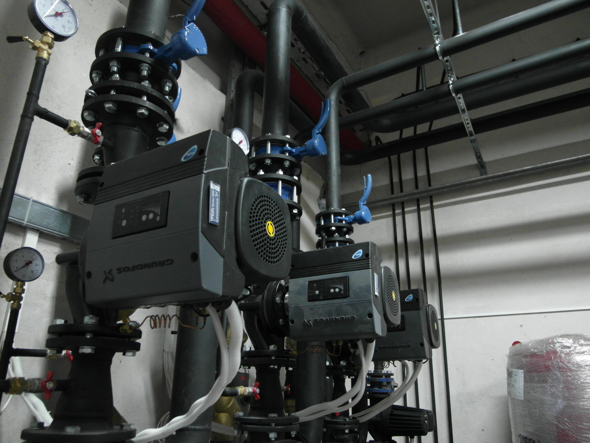 Rząd zainstalowanych pomp obiegowych Grundfos z manometrami i zaworami odcinającymi, połączonych rurami w kotłowni.