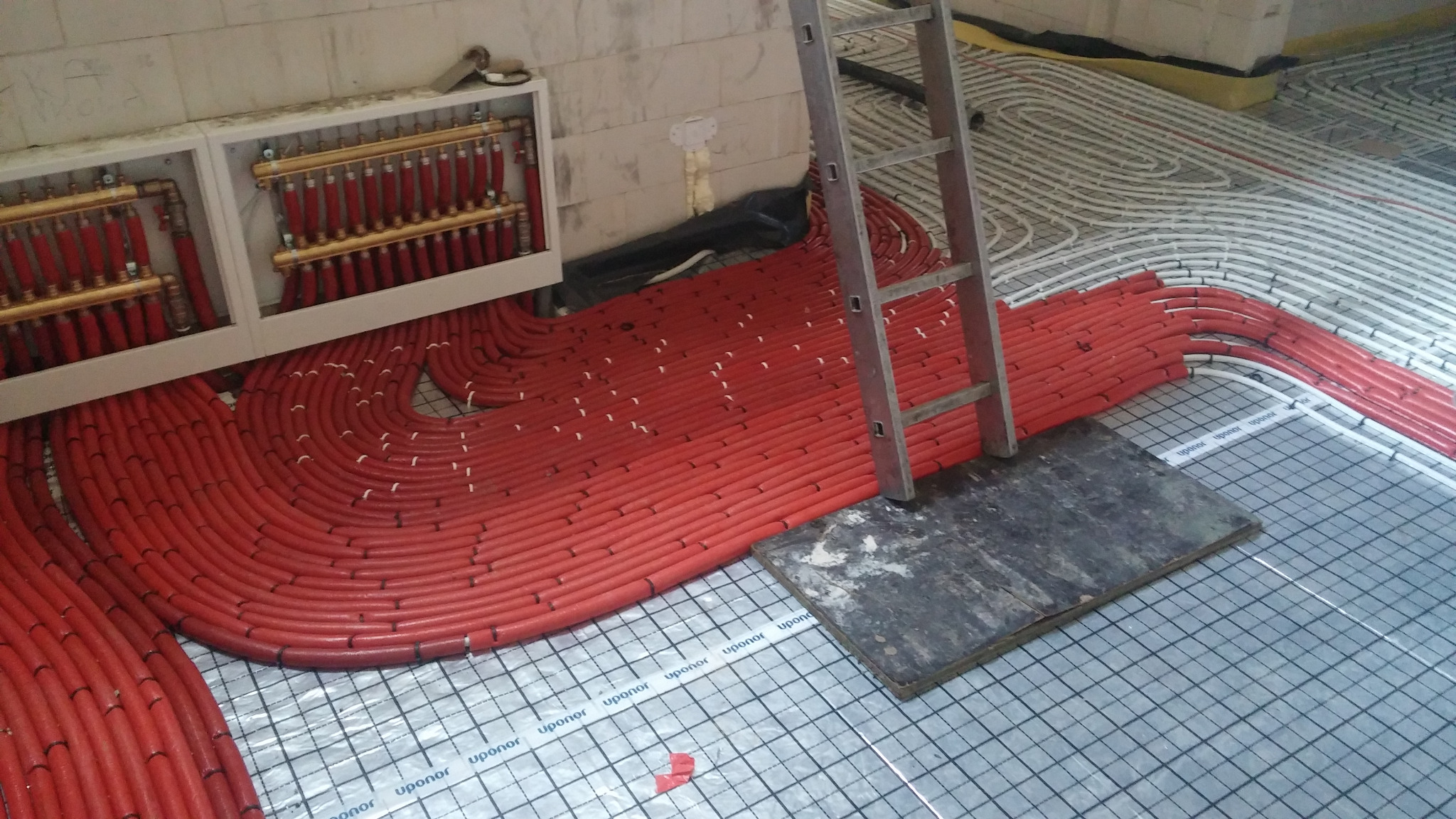 Rozłożone czerwone rury ogrzewania podłogowego na siatce z folią aluminiową, widoczny rozdzielacz w szafce na ścianie i drabina oparta o podłoże.