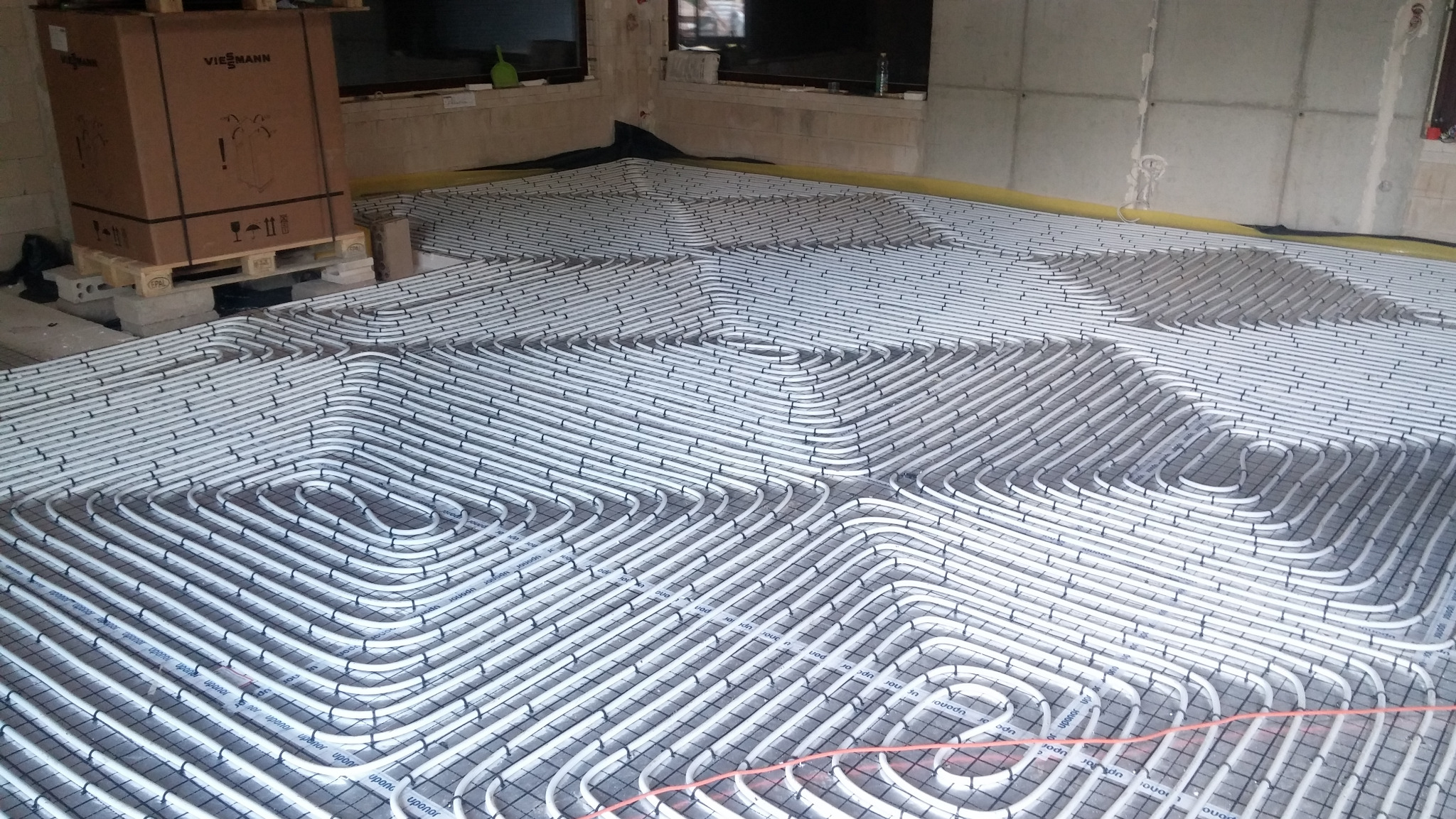Białe rury grzewcze Upor ułożone spiralnie na czarnej siatce w surowym pomieszczeniu, widoczny kartonowy kontener marki Viessmann na palecie w tle.