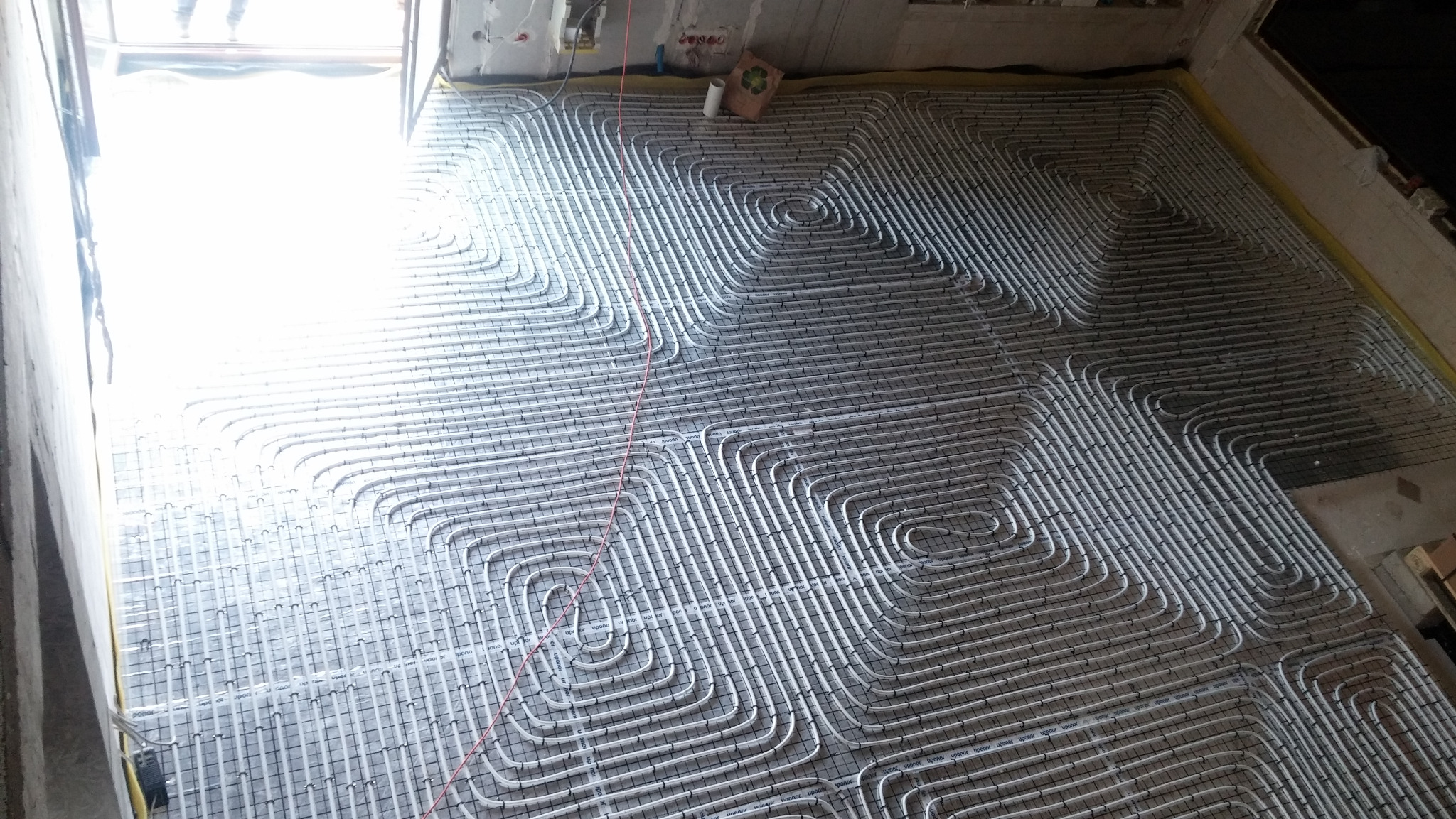 Białe rurki grzewcze ułożone spiralnie na podłodze z siatką montażową, widoczne przygotowania do wylewki, jasne światło wpadające przez okno.
