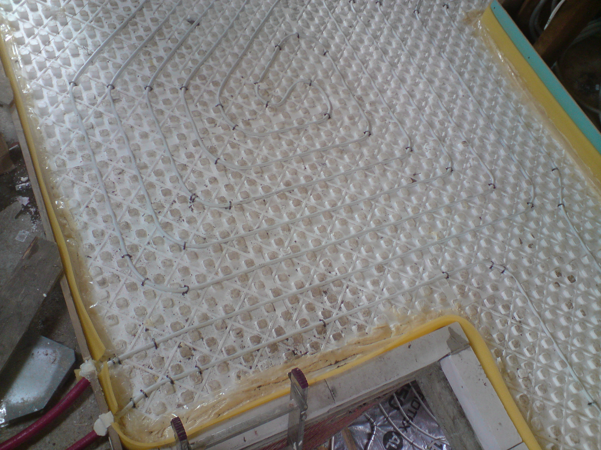 Białe rurki grzewcze rozłożone na podkładzie z siatki, przygotowane do zalania wylewką, widoczne fragmenty izolacji na obrzeżach oraz narzędzia pracy.