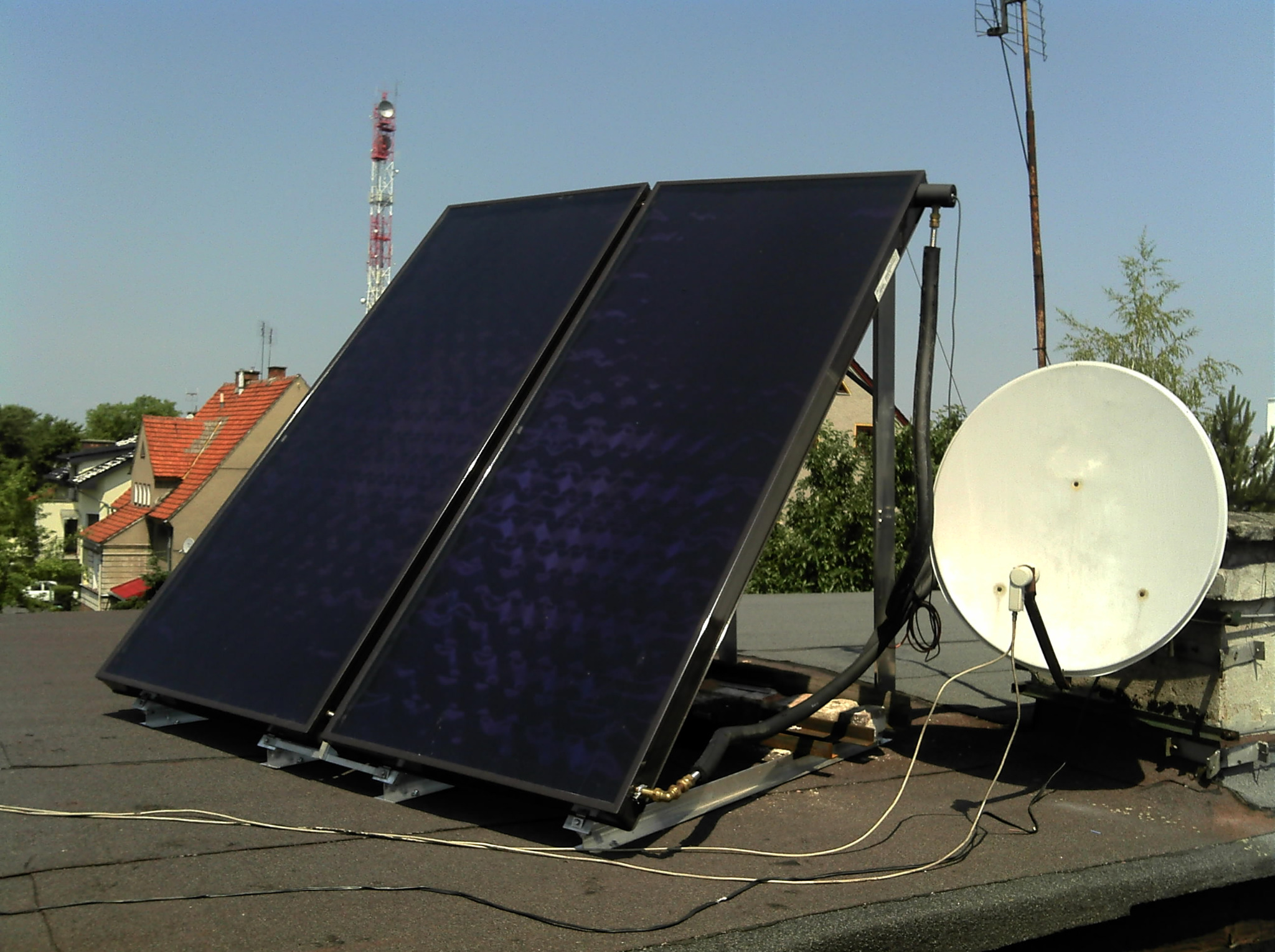 Dwa panele słoneczne zamontowane na dachu budynku z widoczną anteną satelitarną i kominem w tle, w słoneczny dzień.