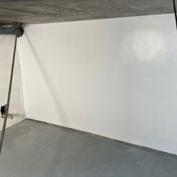 IPA construction - Świeżo pomalowane na biało ściany w pomieszczeniu w trakcie remontu, widoczne szare rury pod sufitem i betonowa podłoga, niedokończone gniazdko elektryczne.