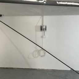 IPA construction - Świeżo wyszpachlowana ściana z widocznymi przewodami elektrycznymi, rurami i elementami instalacji, podparta metalową rozpórką.