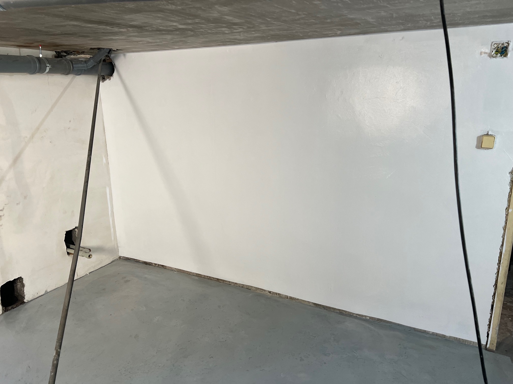Świeżo pomalowane na biało ściany w pomieszczeniu w trakcie remontu, widoczne szare rury pod sufitem i betonowa podłoga, niedokończone gniazdko elektryczne.