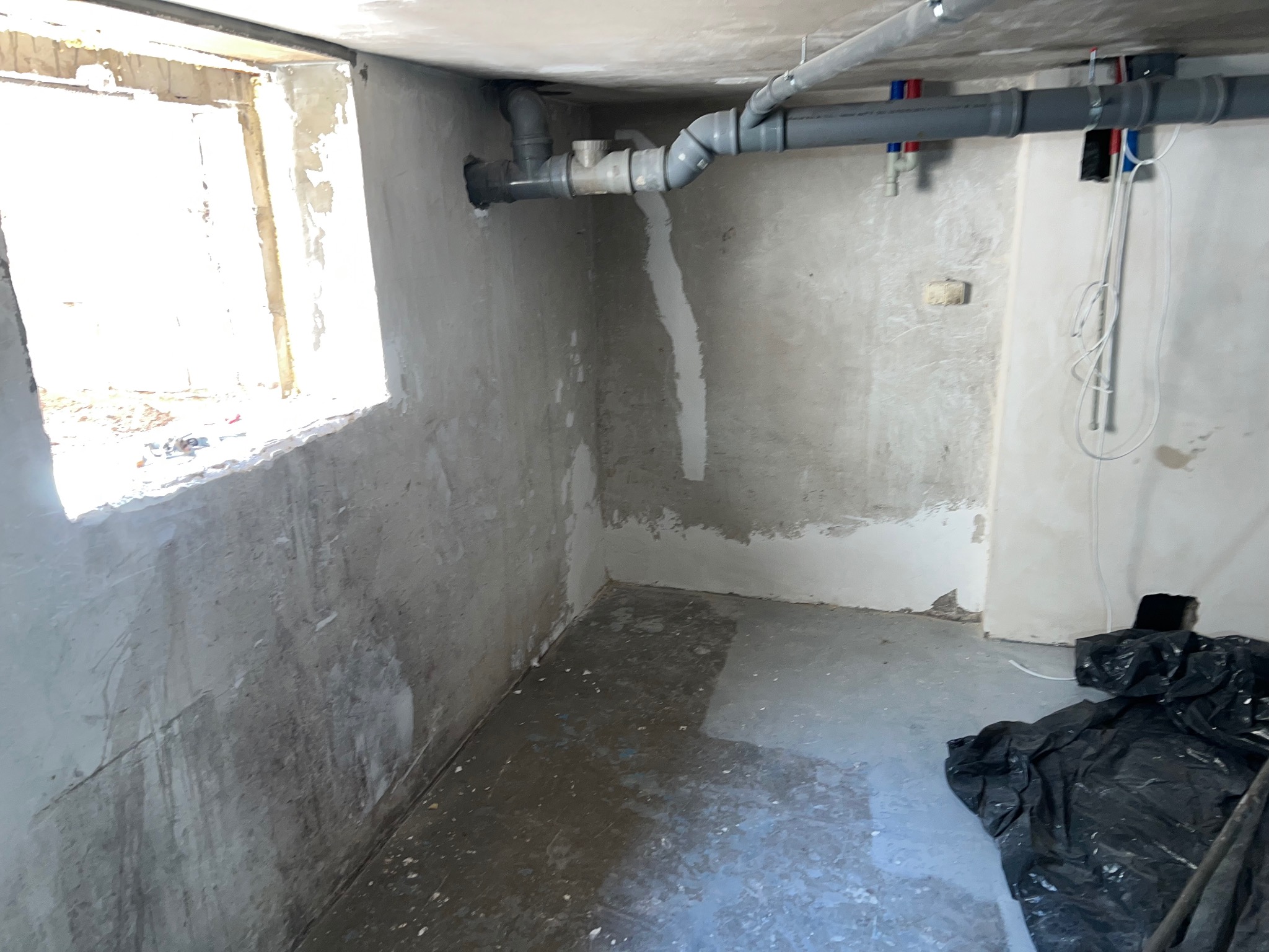 Przygotowanie do remontu w surowym pomieszczeniu z oknem: częściowo wyszpachlowane ściany, rury pod sufitem, zabezpieczona podłoga folią malarską.