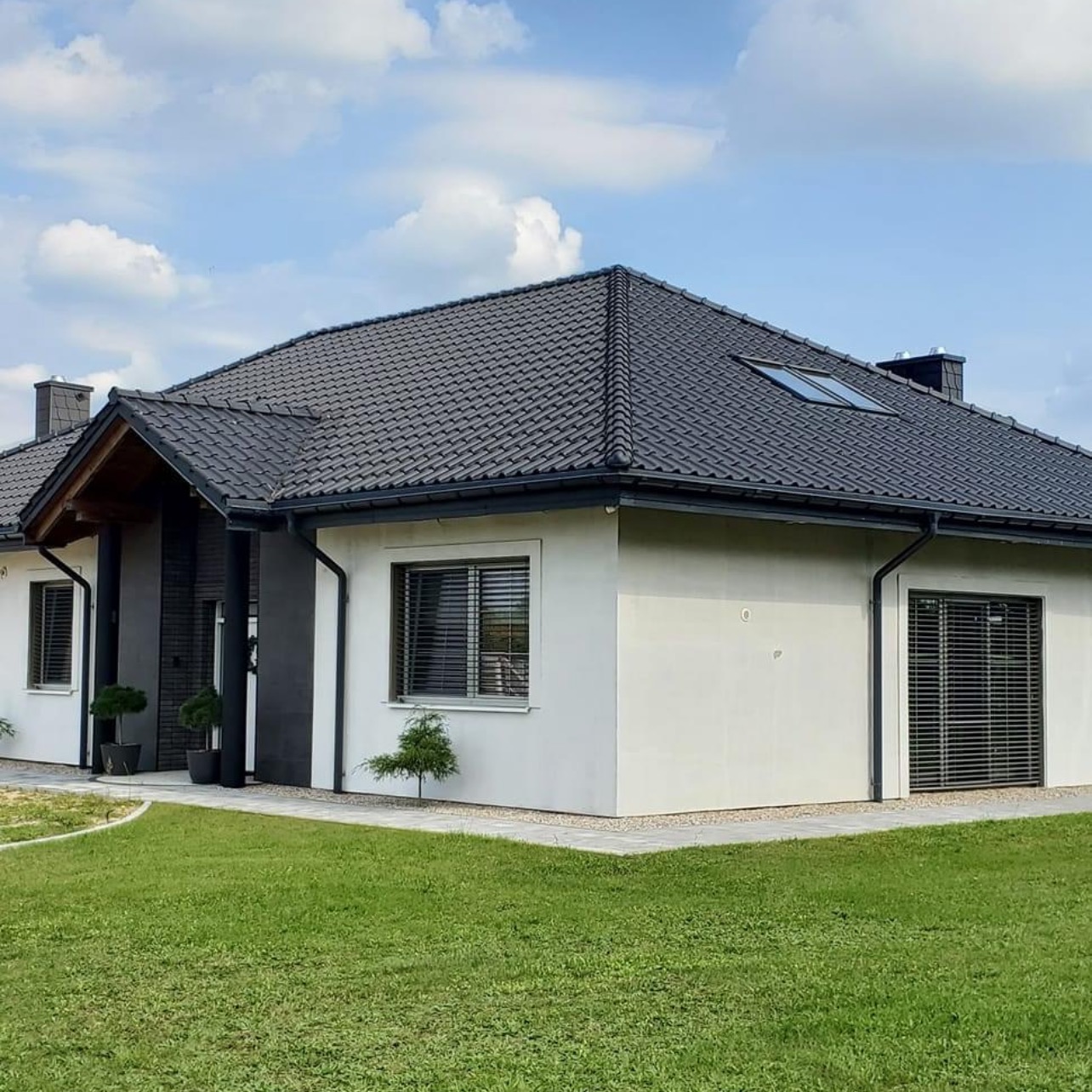 Elewacja domu jednorodzinnego z szarą dachówką, białymi ścianami i ciemnymi elementami wykończeniowymi, widok z trawnika w słoneczny dzień.