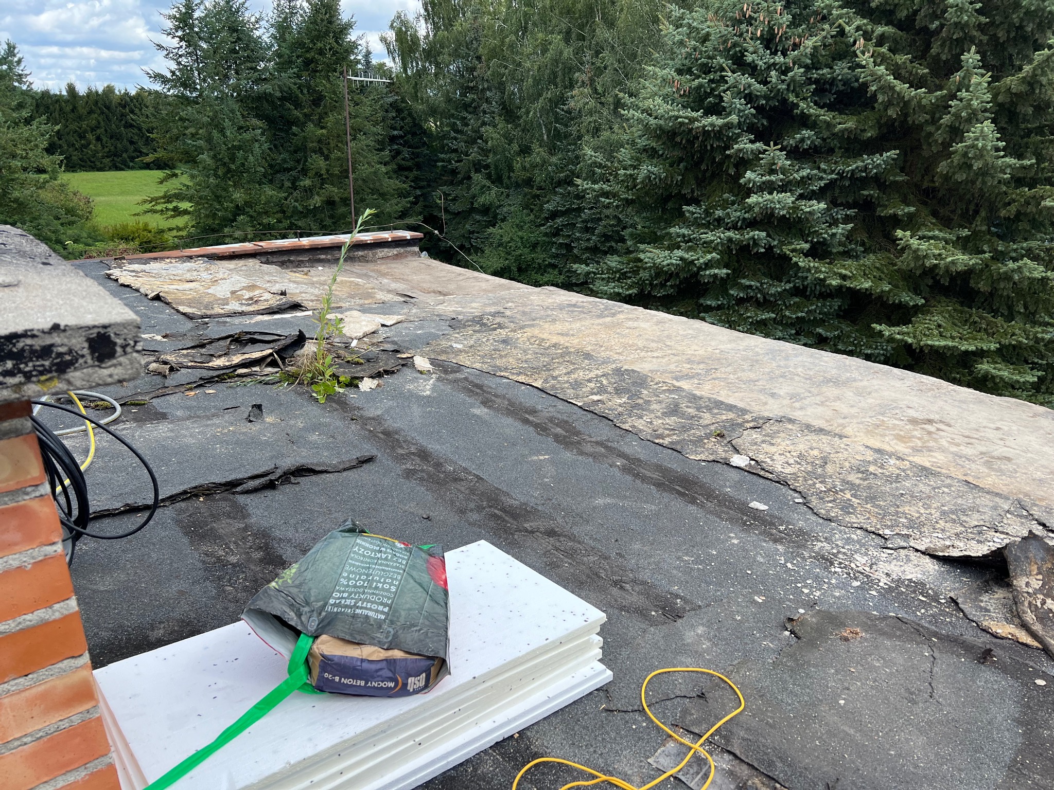 Uszkodzony dach z widocznymi warstwami papy i styropianem przygotowanym do położenia, w tle zieleń drzew.