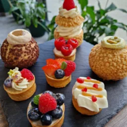 Siedem mini-deserów na czarnym łupkowym talerzu: tartaletki z kremem i owocami (maliny, borówki, truskawki), ptysie z kremem i karmelem, tło rozmyte, rośliny.