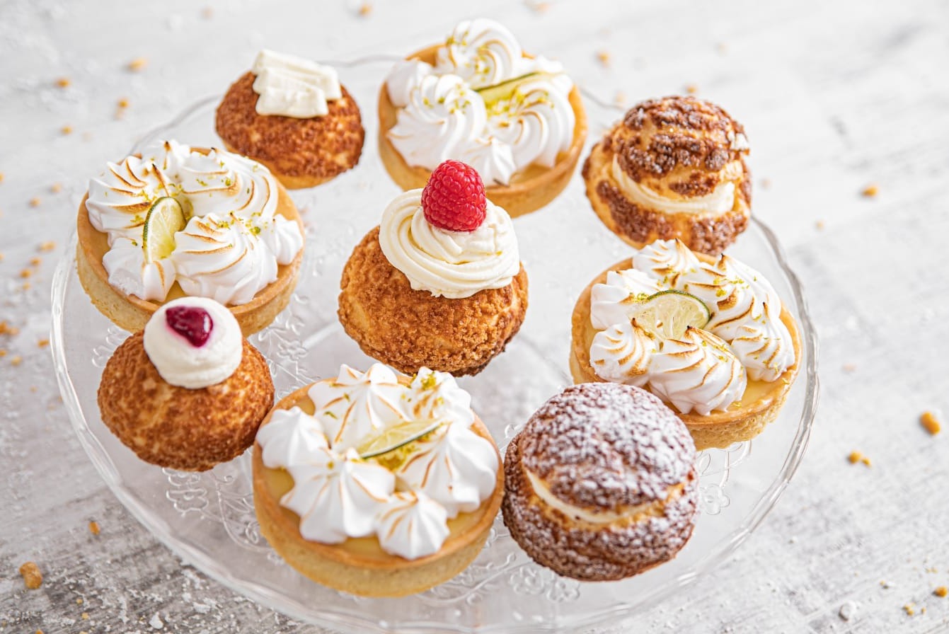 Elegancki talerz z różnorodnymi ciastkami: tartaletki z kremem i limonką, ptysie z kremem i owocami, delikatnie oprószone cukrem pudrem.