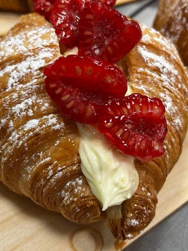 Kremowy rogalik posypany cukrem pudrem, udekorowany kremem i połówkami malin w żelu na drewnianej desce.