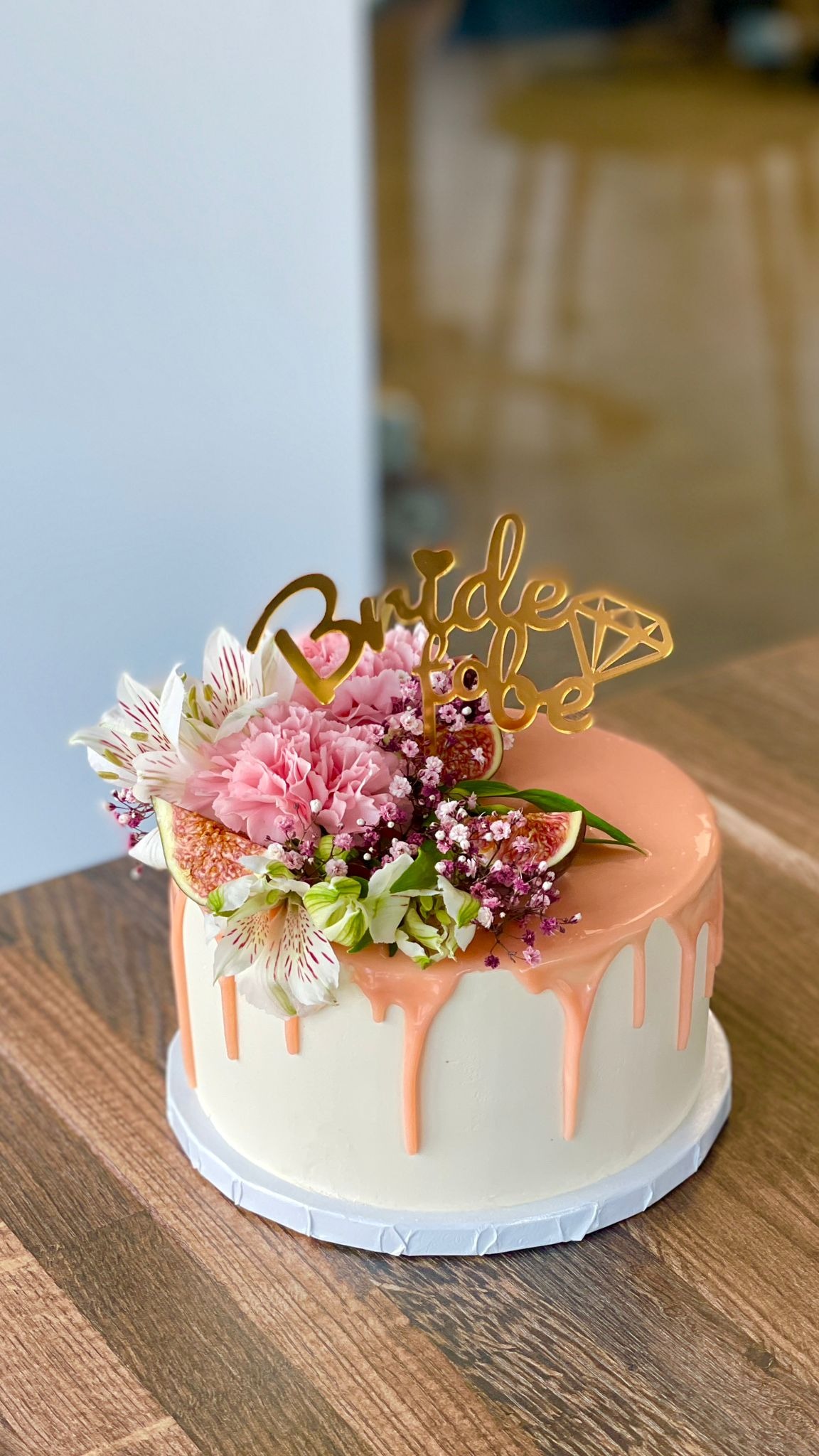 Tort 'Bride to be' z białym kremem, polewą w odcieniach brzoskwini, dekorowany figami, różowymi goździkami, alstromeriami i drobnymi fioletowymi kwiatami, na drewnianym stole.