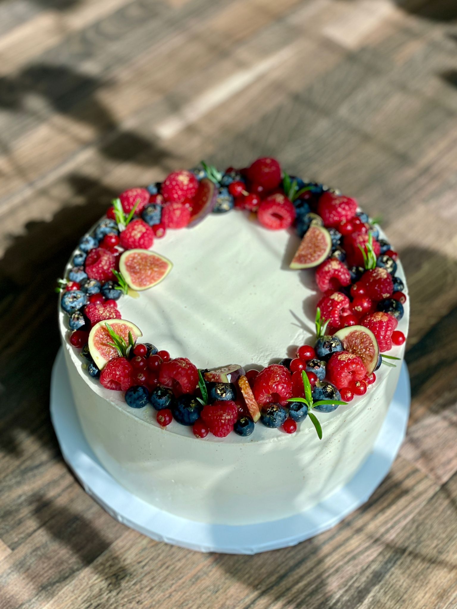 Tort z białym kremem udekorowany wieńcem ze świeżych owoców: malin, borówek, czerwonych porzeczek i plasterków figi, z gałązkami rozmarynu, leżący na drewnianym blacie w słonecznym świetle.