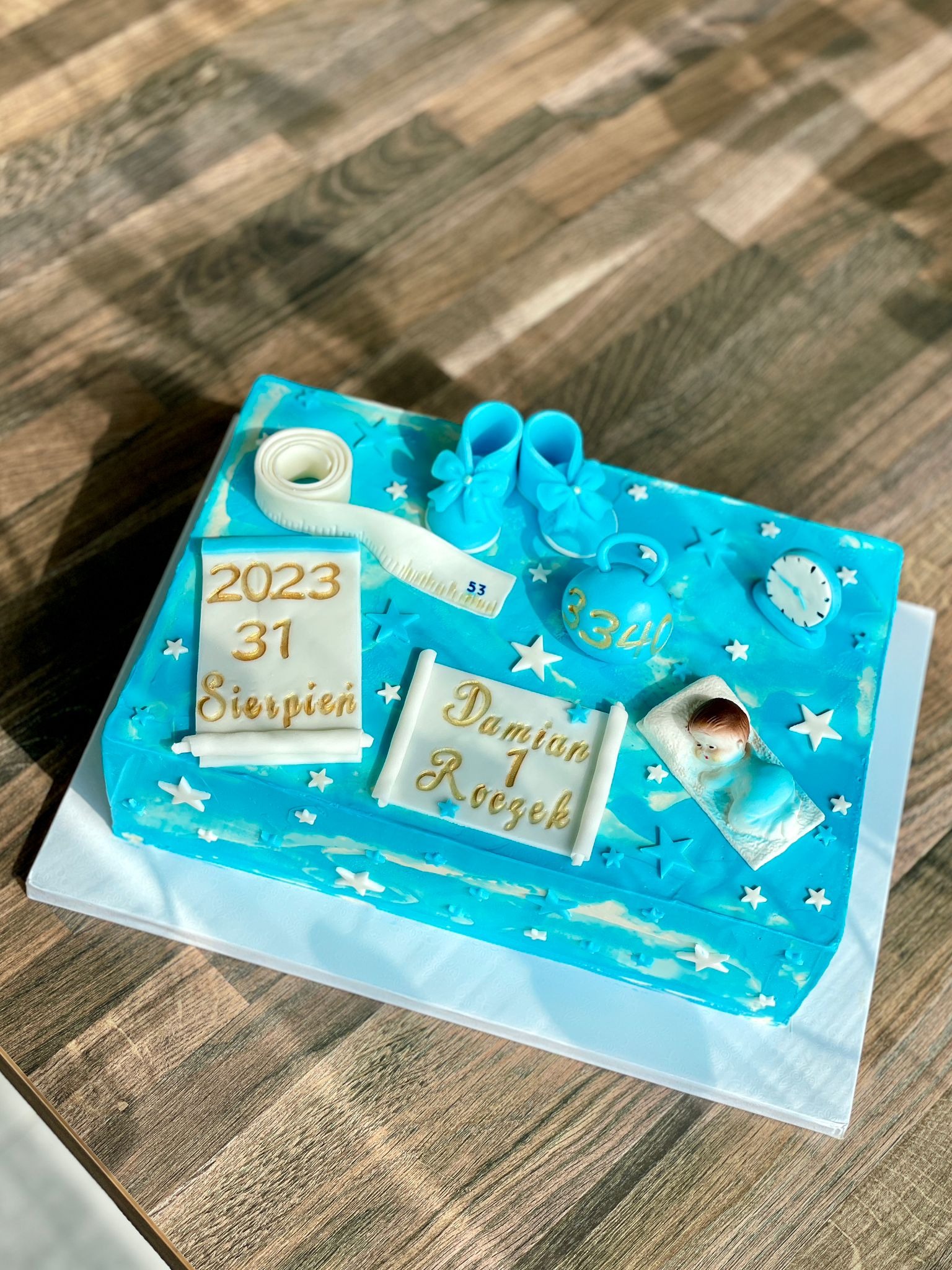 Niebieski tort prostokątny na pierwsze urodziny z dekoracjami: miniaturowe buciki, waga z napisem 3340, zegar, niemowlę na poduszce, kalendarz z datą 31 sierpnia 2023 i napis 'Damian 1 Roczek'
