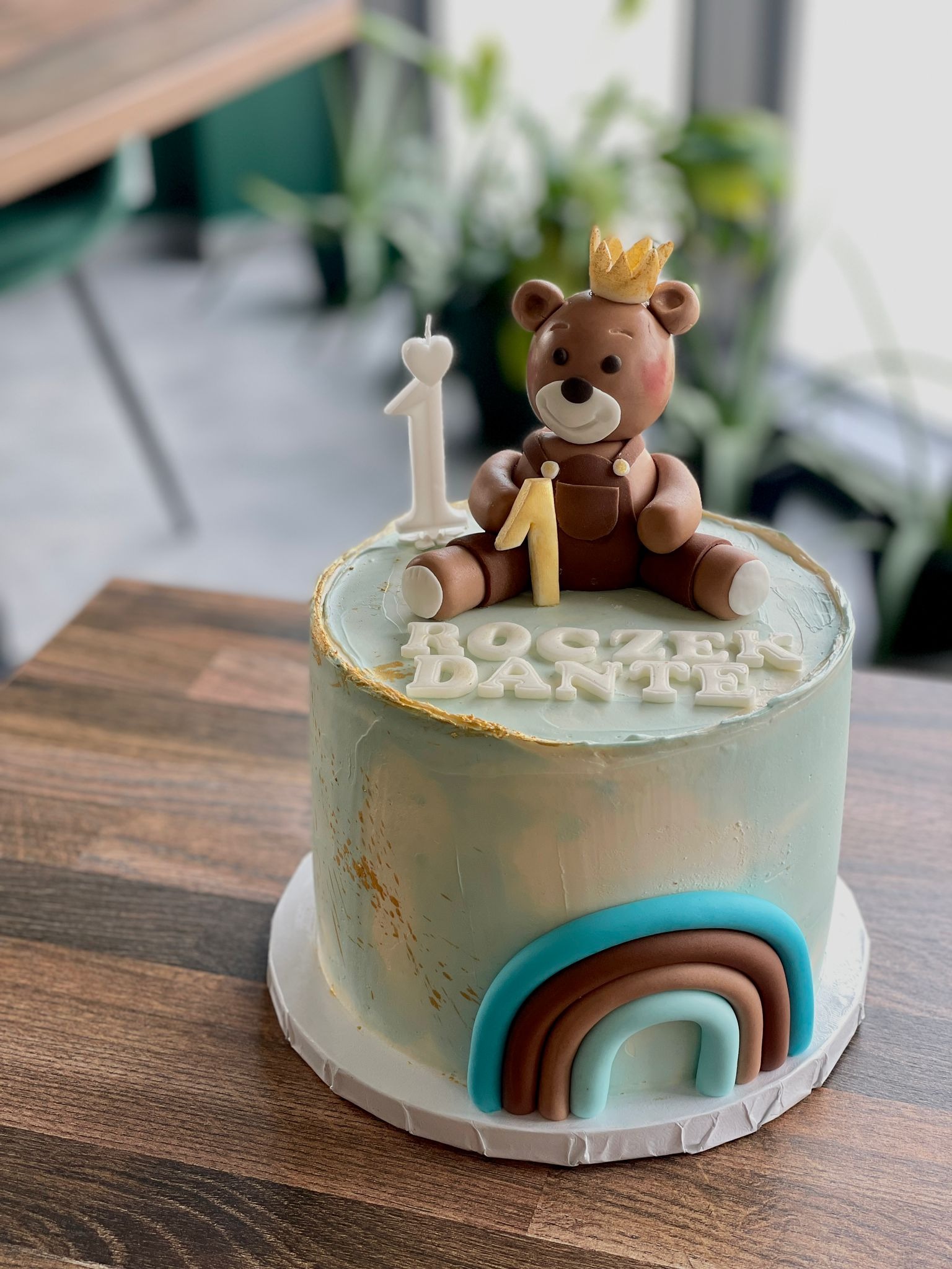 Tort urodzinowy z figurką misia w koronie, świeczką w kształcie cyfry 1, tęczą i napisem 'ROCZEK DANTE'