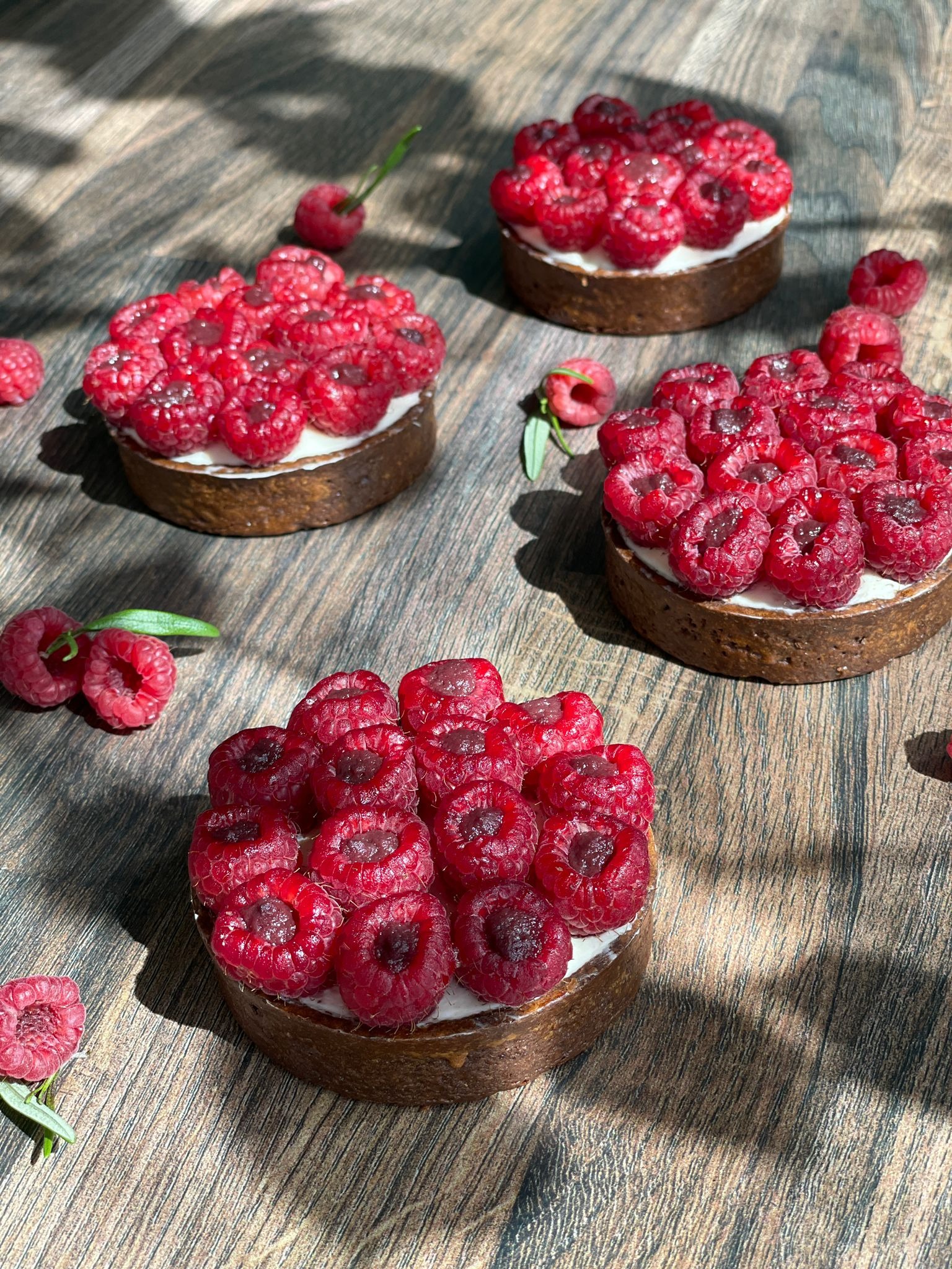 Cztery mini tartaletki z kremem i świeżymi malinami ułożone na drewnianym stole w plamach słonecznego światła.