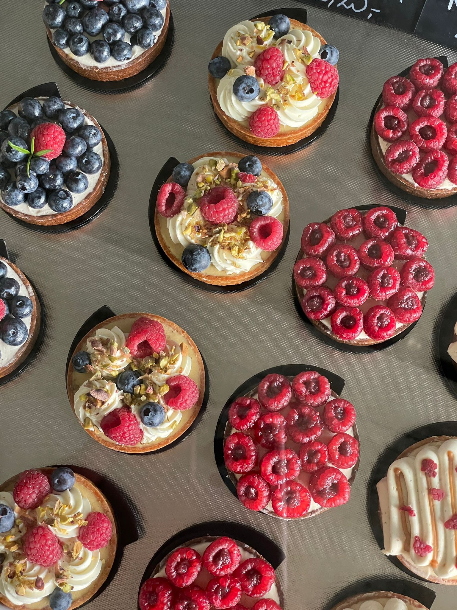 Różnorodne tartaletki z owocami: maliny, borówki, pistacje, na kruchym spodzie z kremowym nadzieniem, ułożone na metalowej powierzchni.