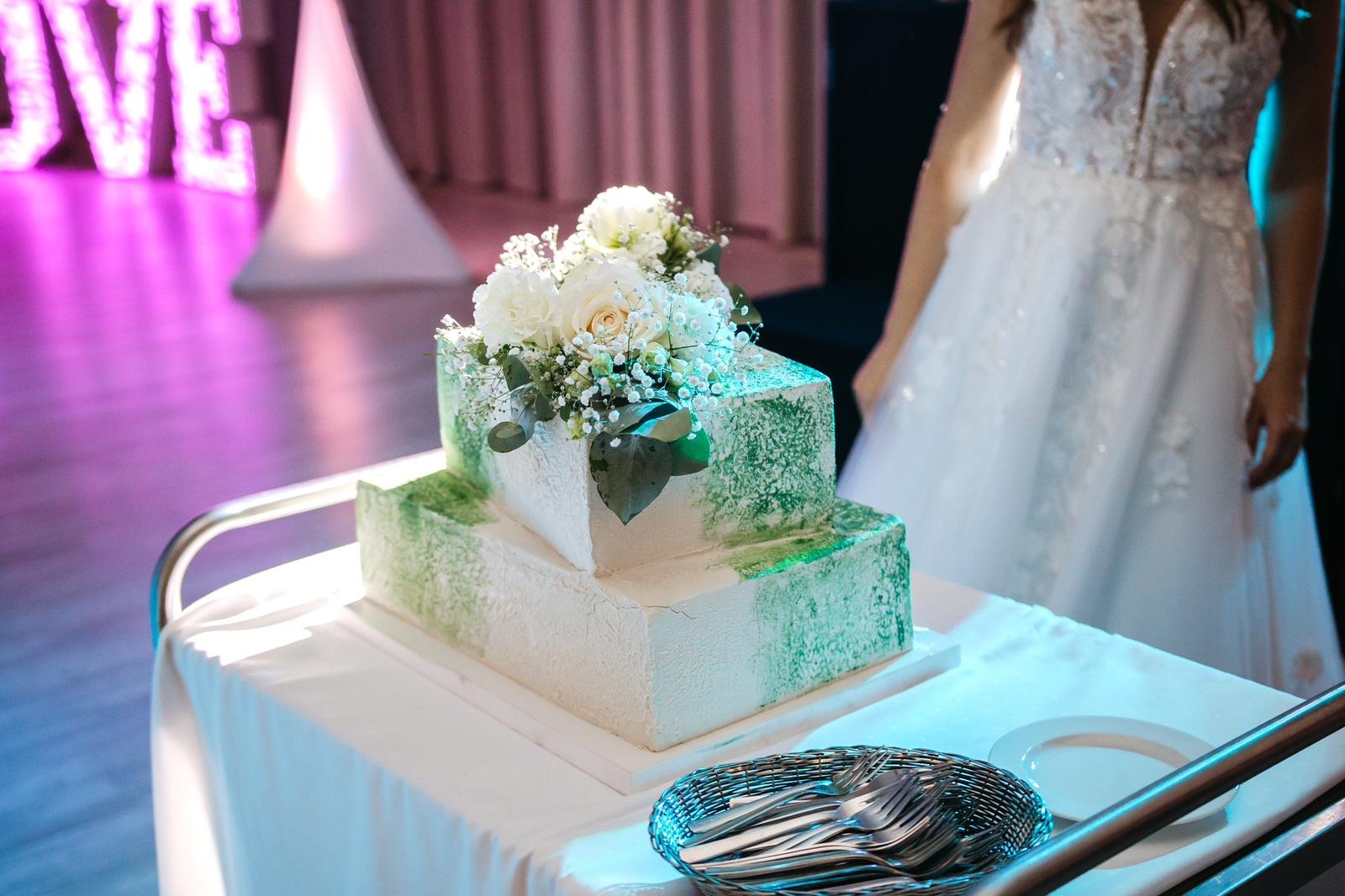 Dwupoziomowy tort weselny w odcieniach bieli i zieleni, udekorowany świeżymi kwiatami, prezentowany w Poznaniu na stole z białym obrusem, obok widoczna suknia panny młodej.
