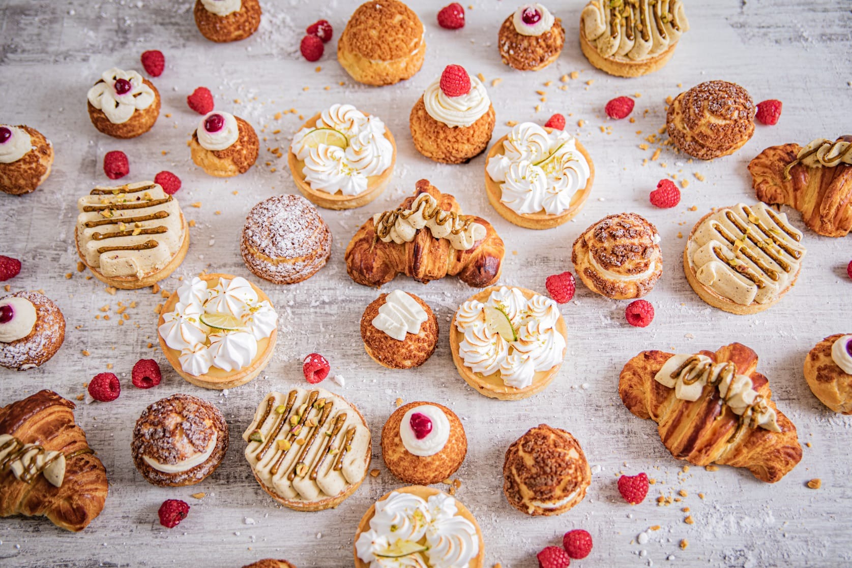 Różnorodne ciastka i croissanty z kremami, owocami i posypkami, ułożone na białym, lekko przetartym drewnianym stole, z rozrzuconymi malinami.