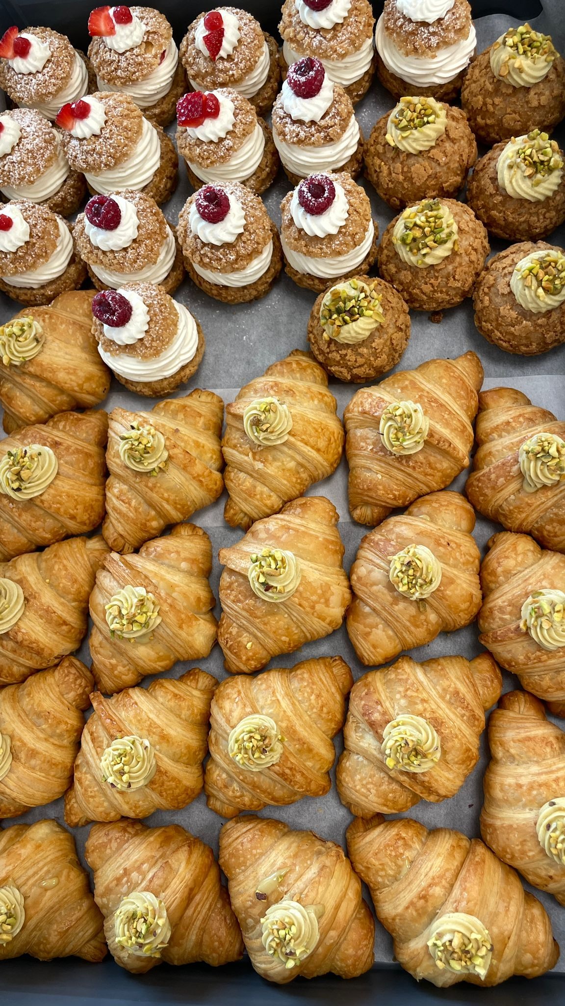 Widok z góry na tacę z różnorodnymi wypiekami: rogaliki francuskie z kremem pistacjowym, ptysie z kremem i malinami oraz ptysie z kremem pistacjowym.