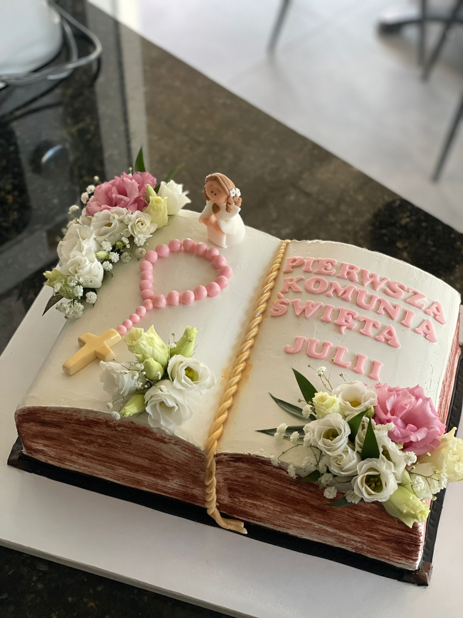 Tort w kształcie otwartej księgi z napisem 'Pierwsza Komunia Święta Julii', ozdobiony kwiatami, różańcem i figurką aniołka.