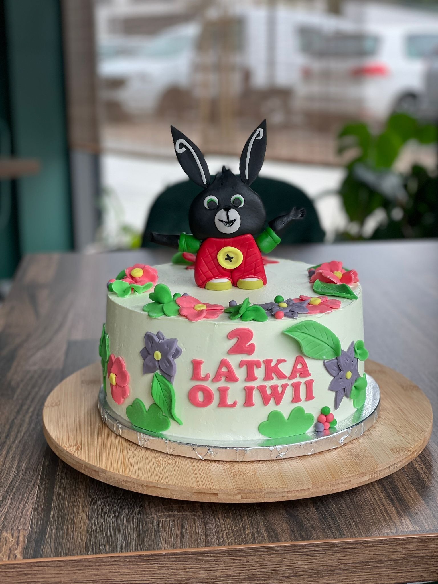 Tort urodzinowy dla dwulatki, z figurką czarnego królika w czerwonym ubranku, otoczony kwiatami z lukru i napisem '2 latka Oliwii', umieszczony na drewnianej paterze.