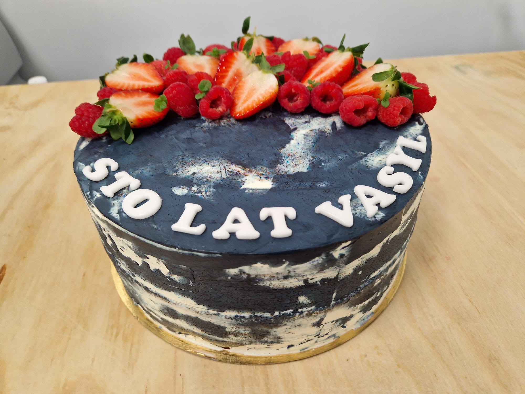 Tort urodzinowy w stylu galaxy z napisem 'Sto lat Vasyl', udekorowany świeżymi truskawkami i malinami, na drewnianym blacie.