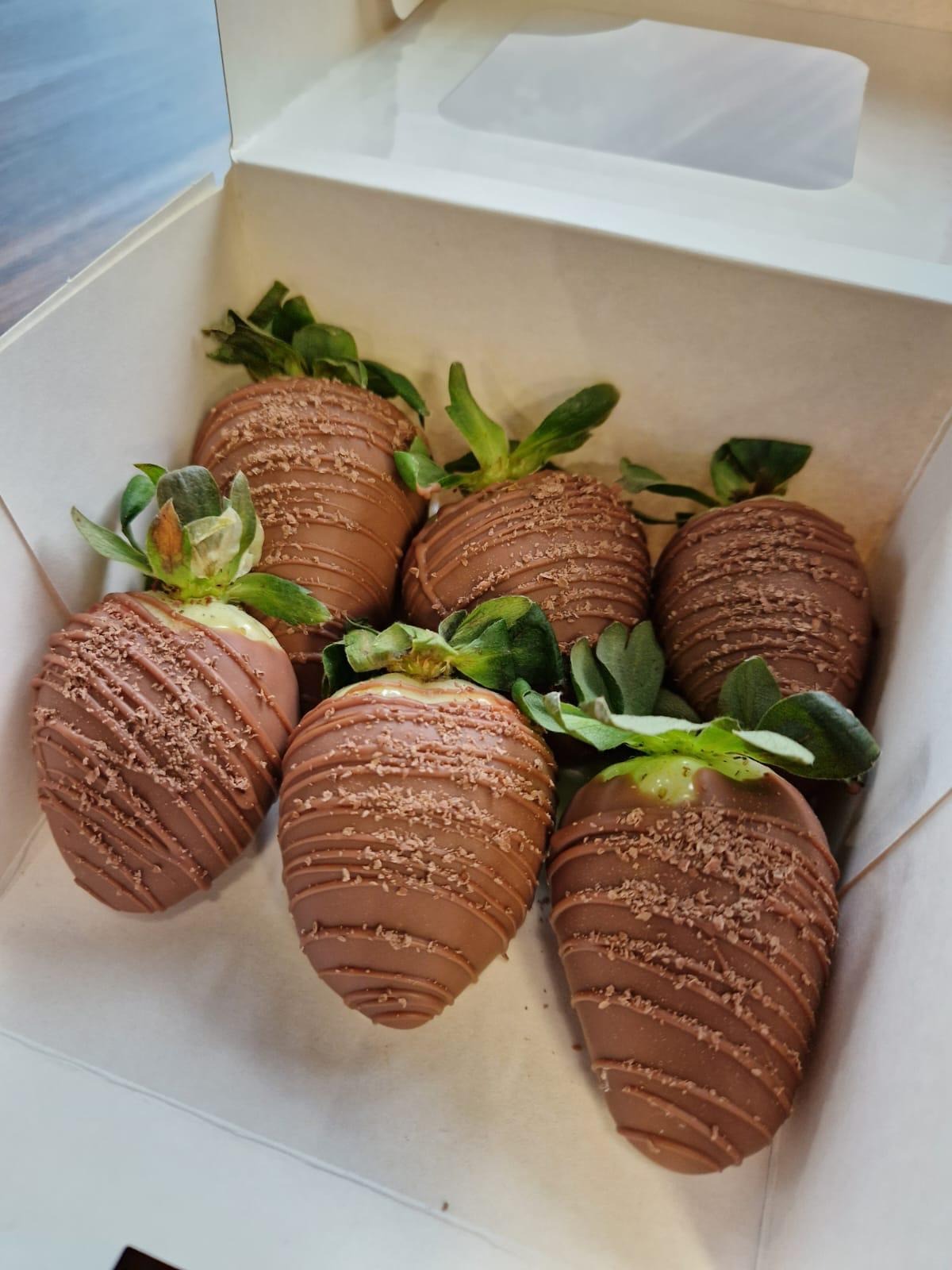 Sześć truskawek w mlecznej czekoladzie, ułożonych w białym kartonowym pudełku z widocznym okienkiem, każda posypana delikatnymi wiórkami czekoladowymi i zielonymi szypułkami.