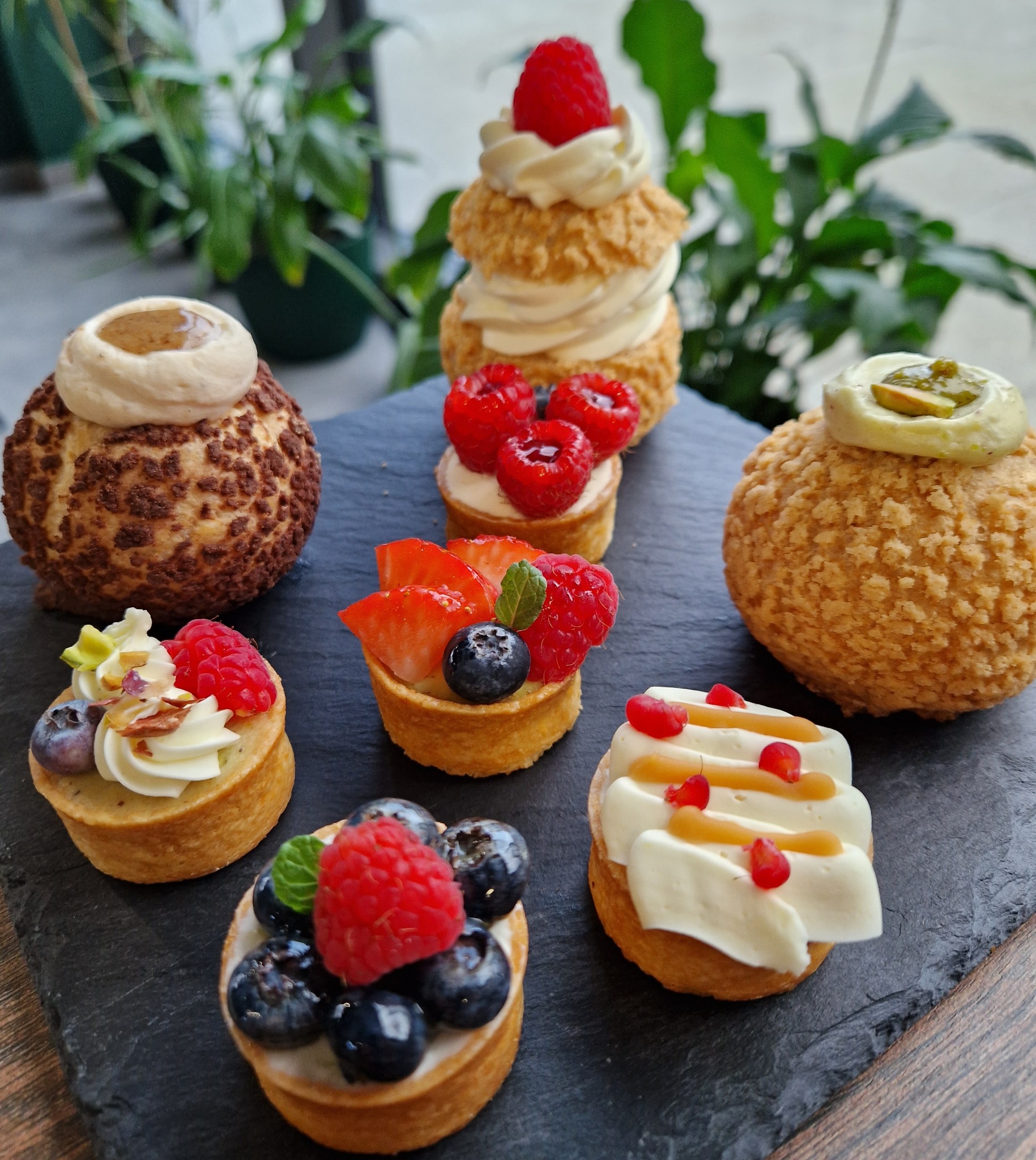 Siedem mini-deserów na czarnym łupkowym talerzu: tartaletki z kremem i owocami (maliny, borówki, truskawki), ptysie z kremem i karmelem, tło rozmyte, rośliny.