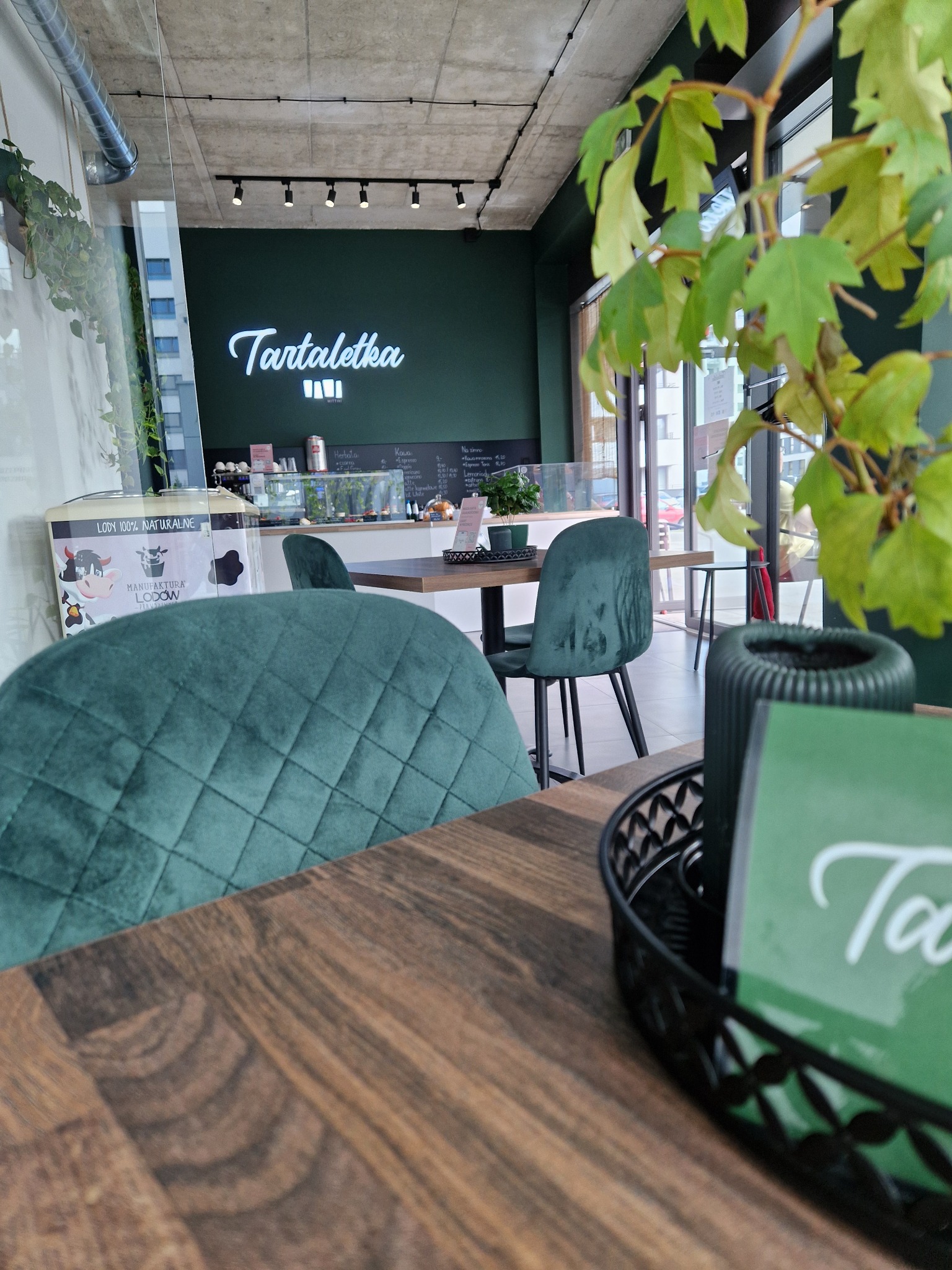 Wnętrze kawiarni z zielonymi, welurowymi krzesłami, drewnianymi stolikami, roślinami w doniczkach i neonowym napisem 'Tartaletka' na ścianie. W tle witryna z deserami i lodami.