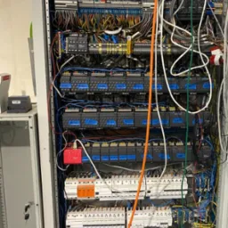 Otwarta szafa sterownicza z widocznymi elementami elektrycznymi, w tym licznymi przewodami w różnych kolorach, modułami sterującymi i zabezpieczeniami; obok szafy widoczny kluczyk i fragment...