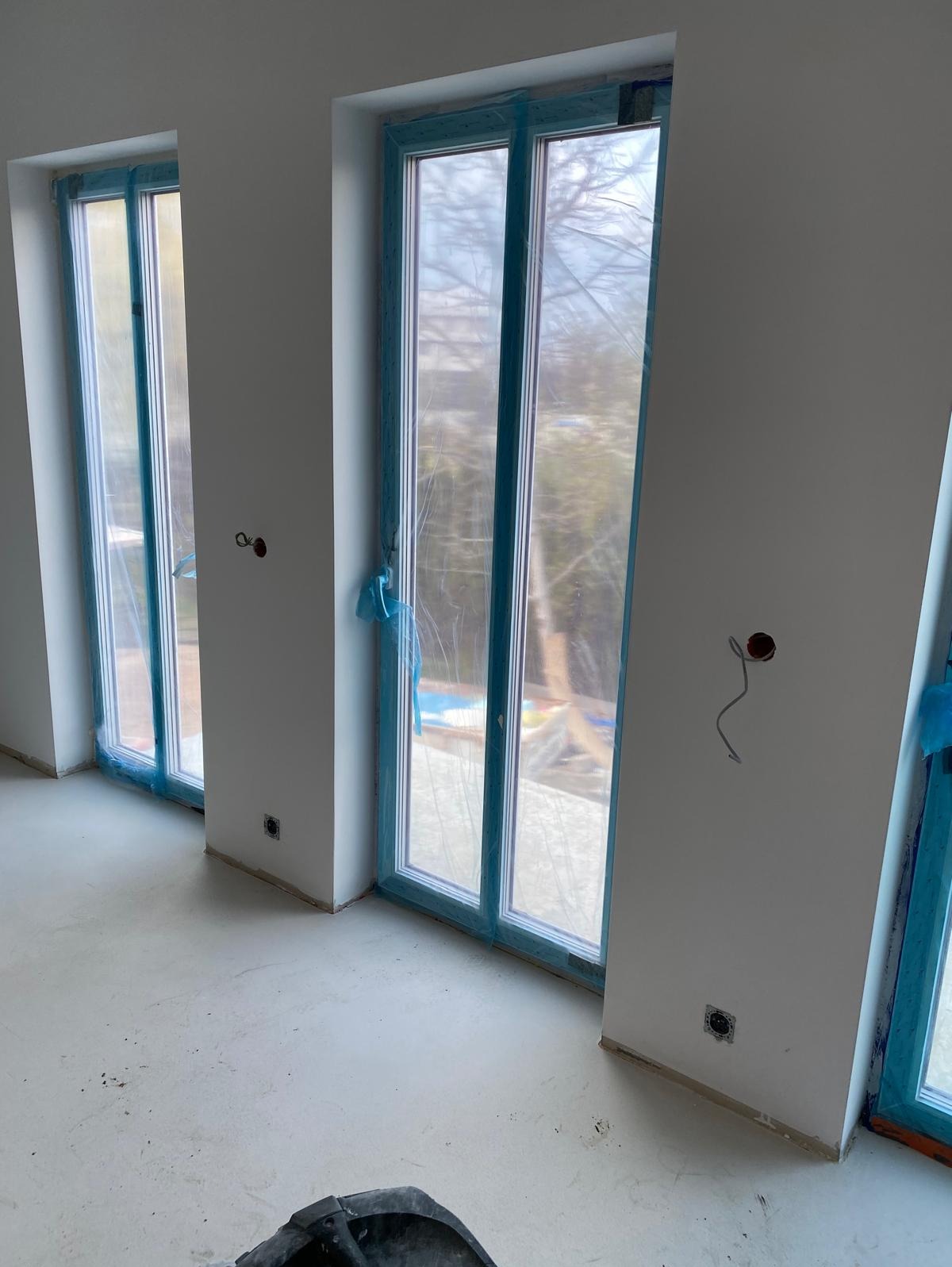 Wnętrze w trakcie remontu: dwa okna zabezpieczone folią ochronną, widoczne wystające przewody elektryczne, surowy beton na podłodze, wiadro w rogu.