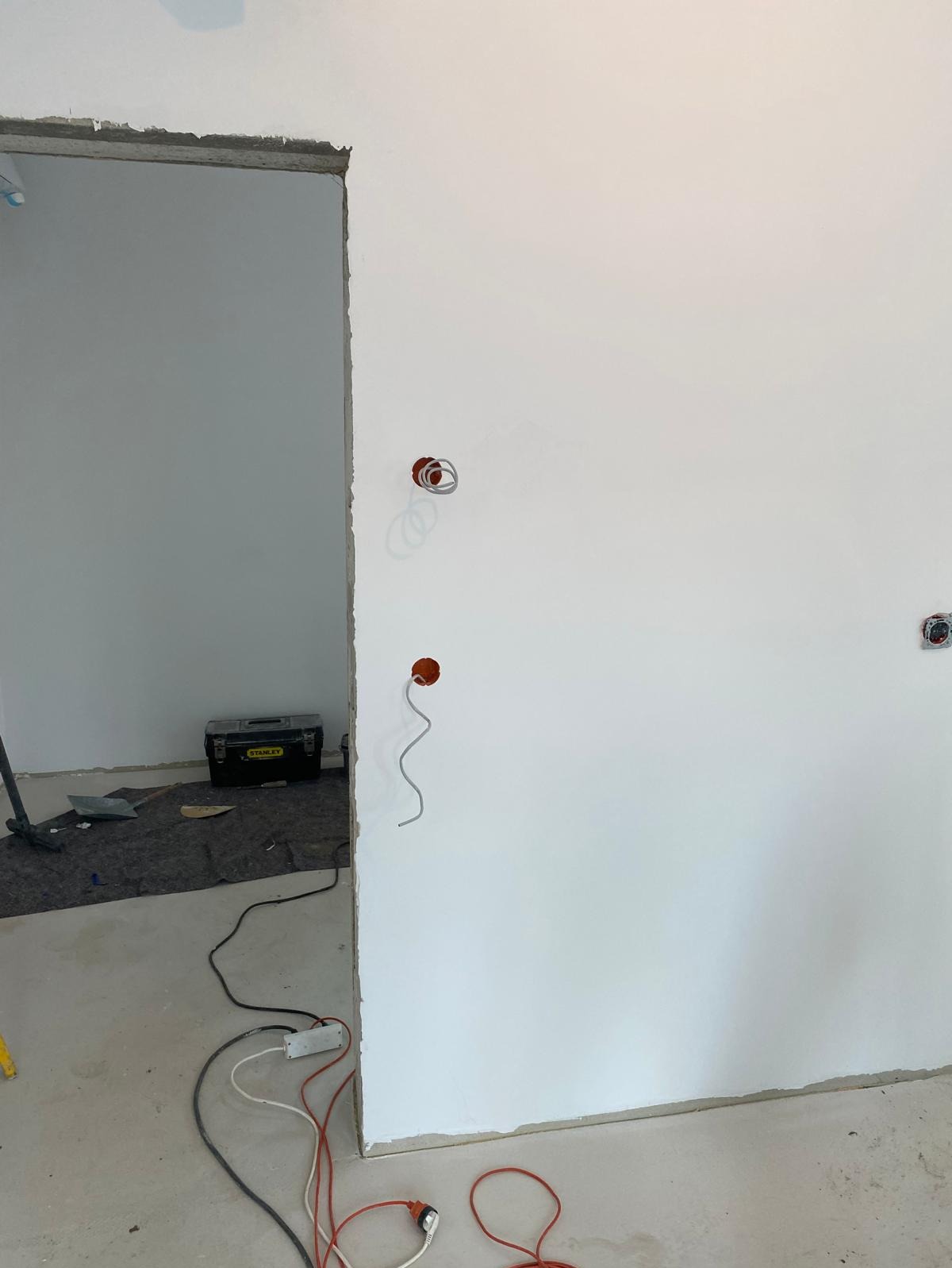 Wnętrze w trakcie remontu: biała ściana z trzema otworami pod gniazdka elektryczne, z których zwisają kable. Widoczna skrzynka narzędziowa Stanley w tle i kable zasilające na podłodze.